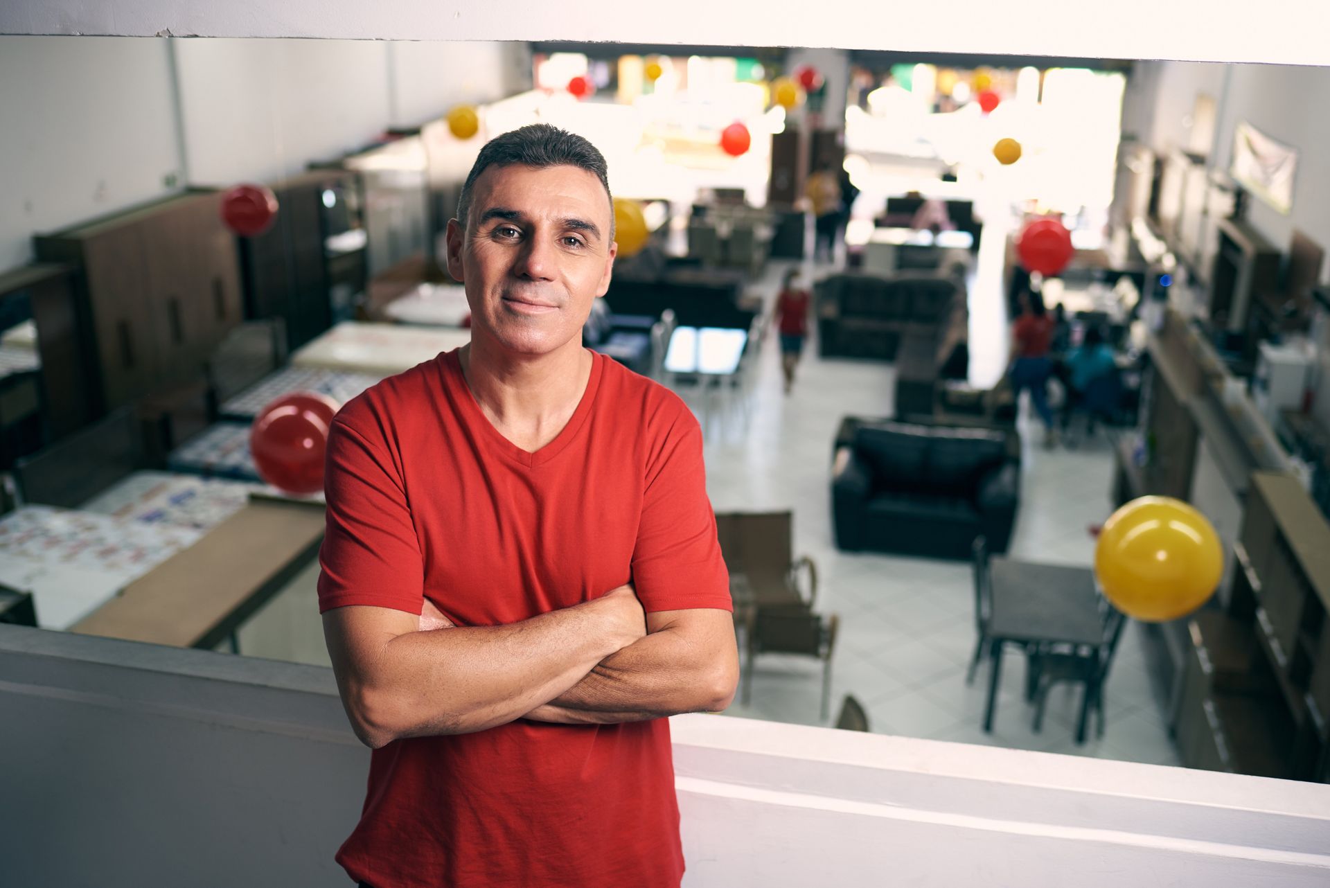 Um homem de camisa vermelha está em uma loja de móveis com os braços cruzados.