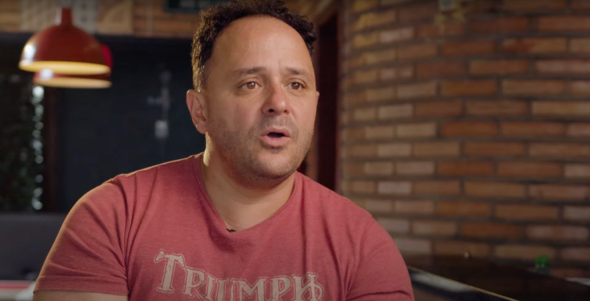 A man in a red t-shirt is sitting at a table in front of a brick wall.