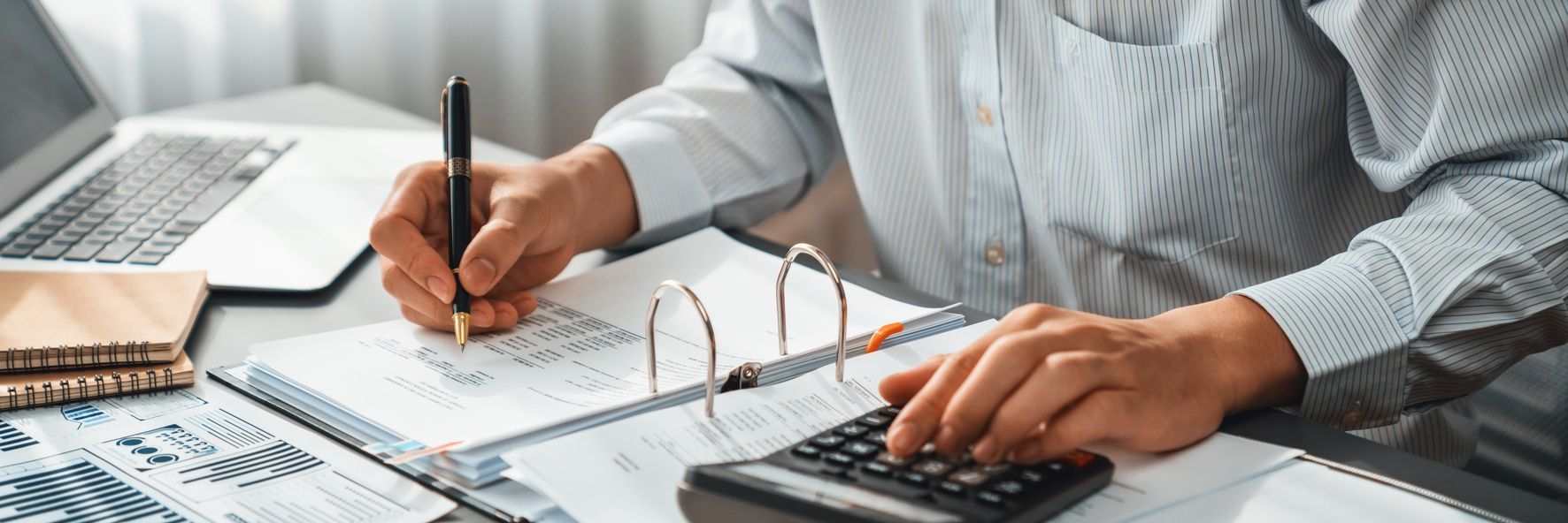 Person working on accounting with pen, calculator, and laptop on desk.