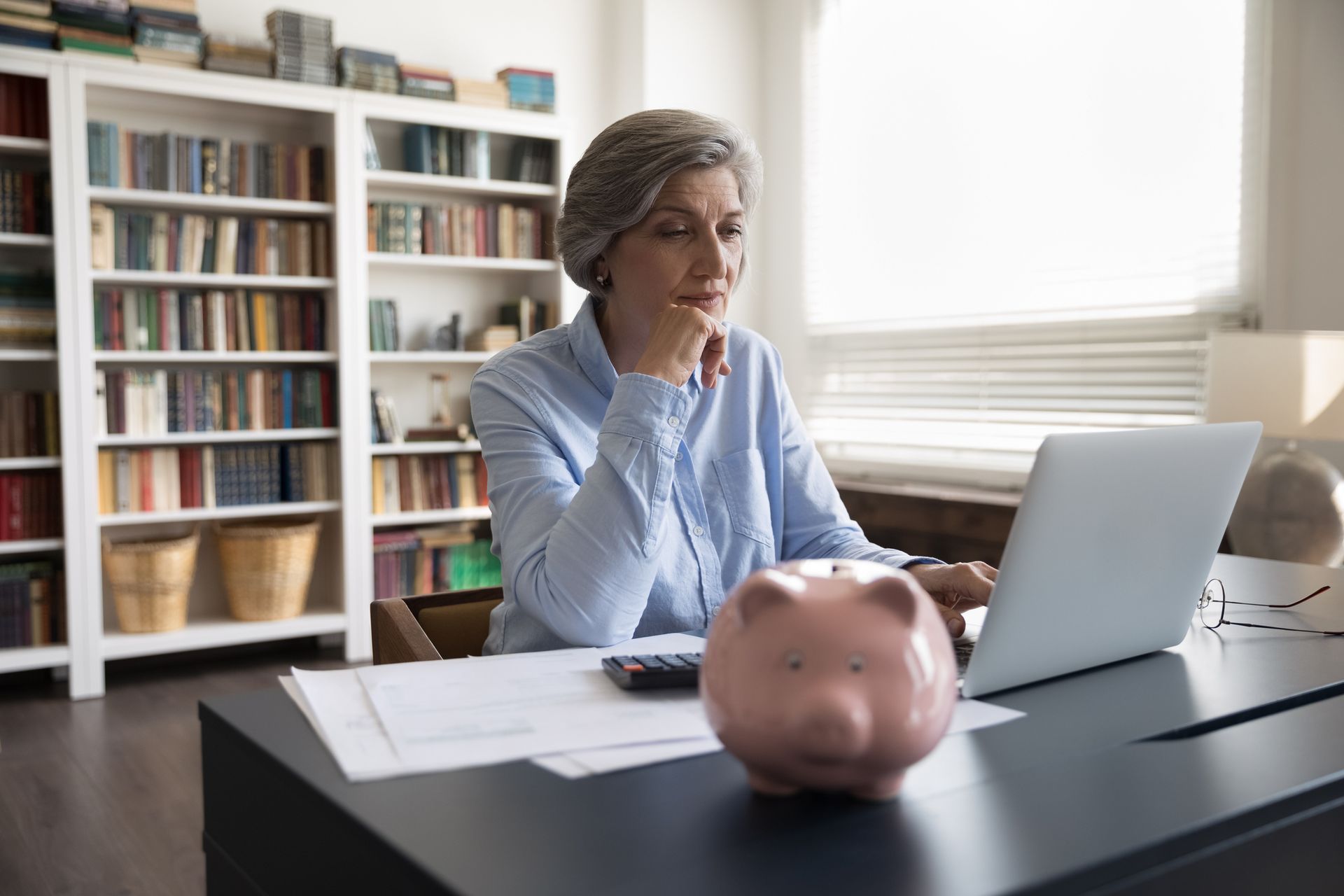 Woman with short gray hair using laptop at desk with piggy bank and bookshelves.