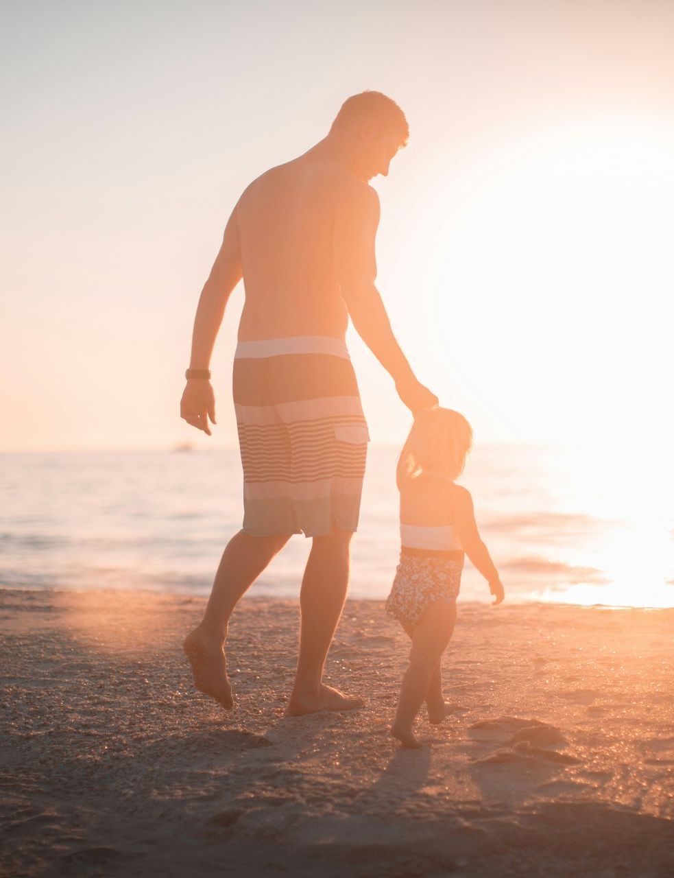 A man and a little girl are walking on the beach at sunset.