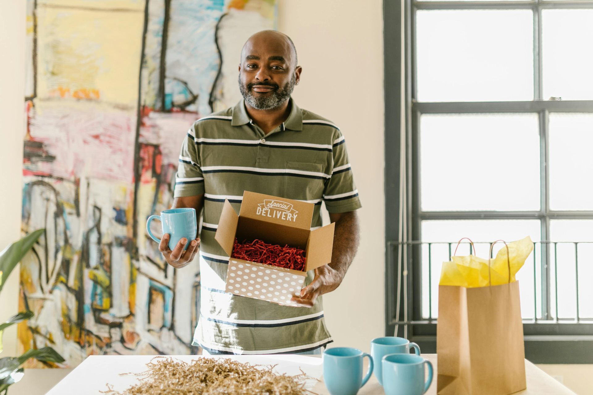 A man is standing in front of a table holding a box and a cup.