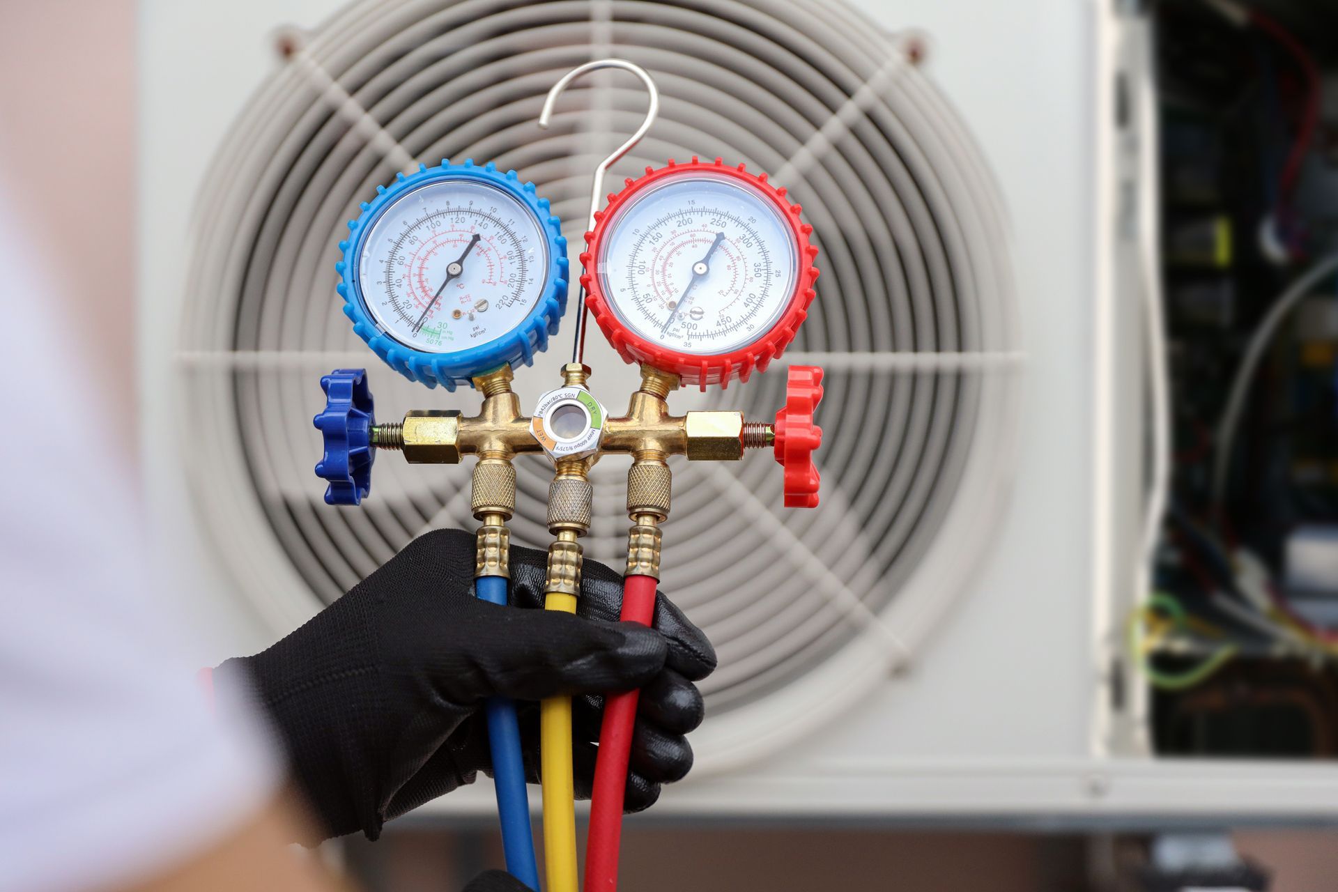 Technician holding HVAC manifold gauges with blue and red dials in front of an outdoor unit fan