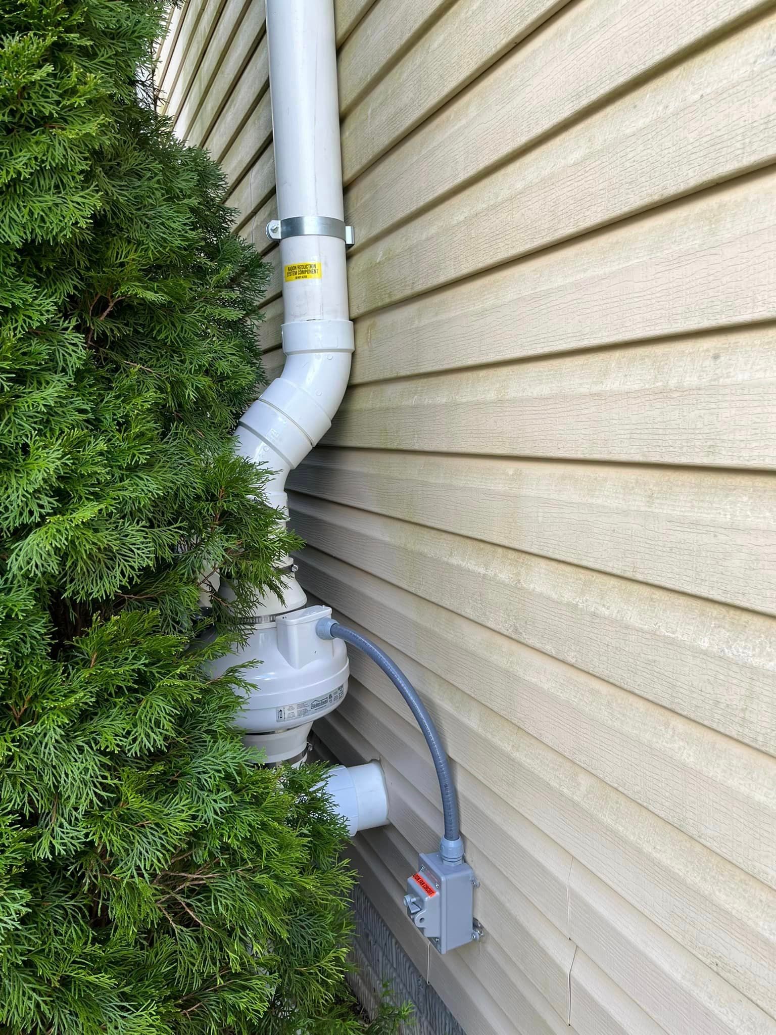 A white pipe is attached to the side of a house next to a bush.