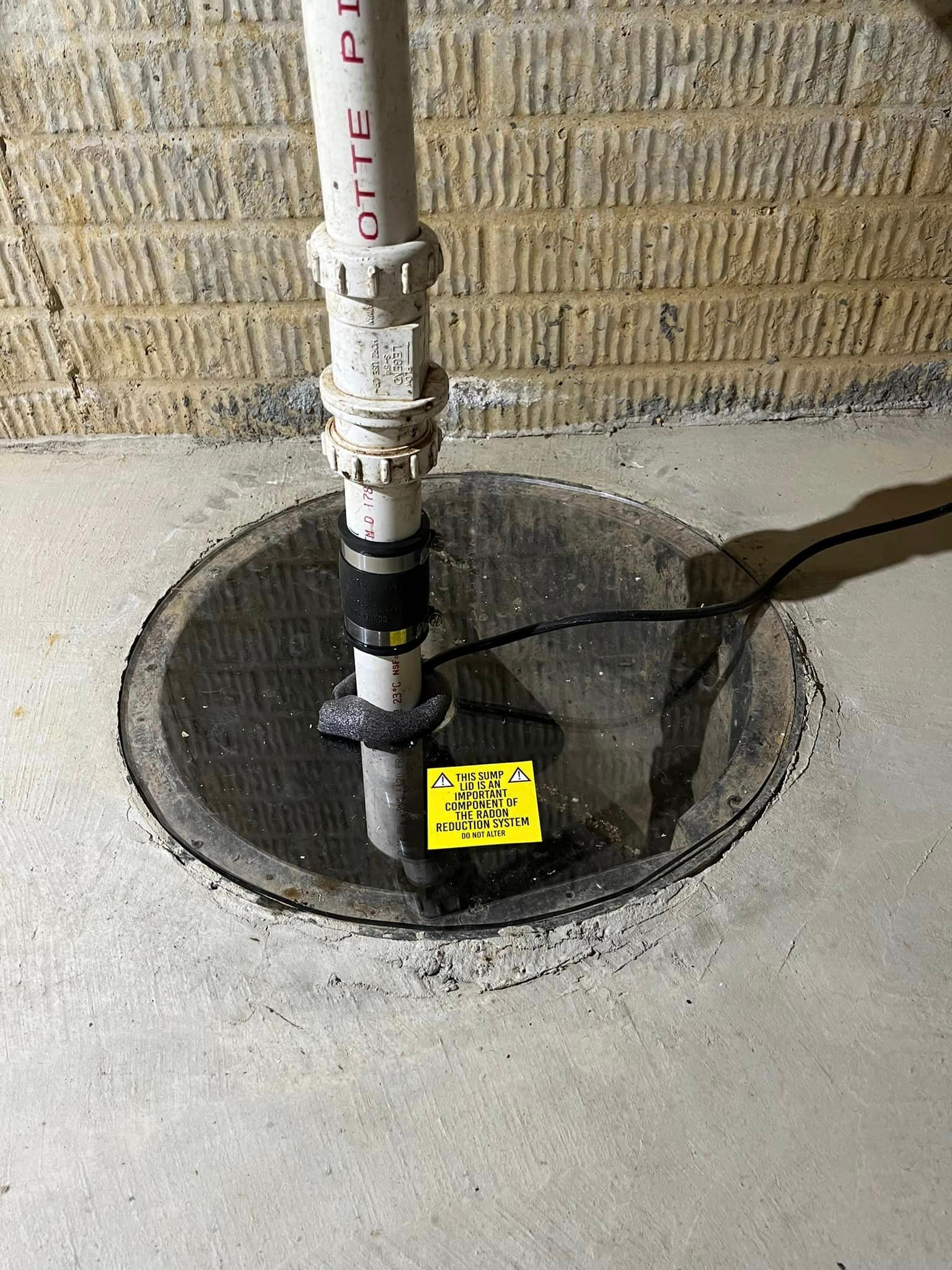 A sump pump is sitting in a basement next to a pipe.