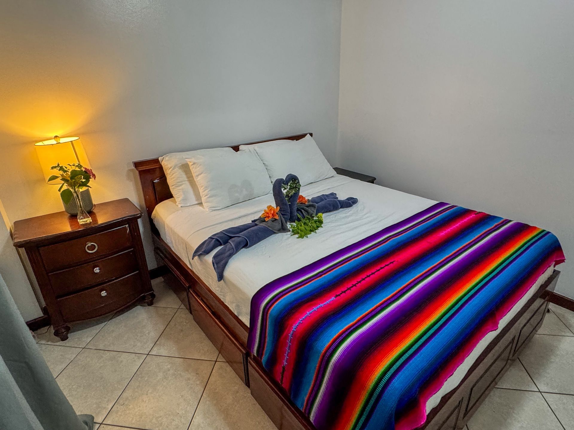 A bedroom with a bed , nightstand and lamp . the bed has a colorful blanket on it.