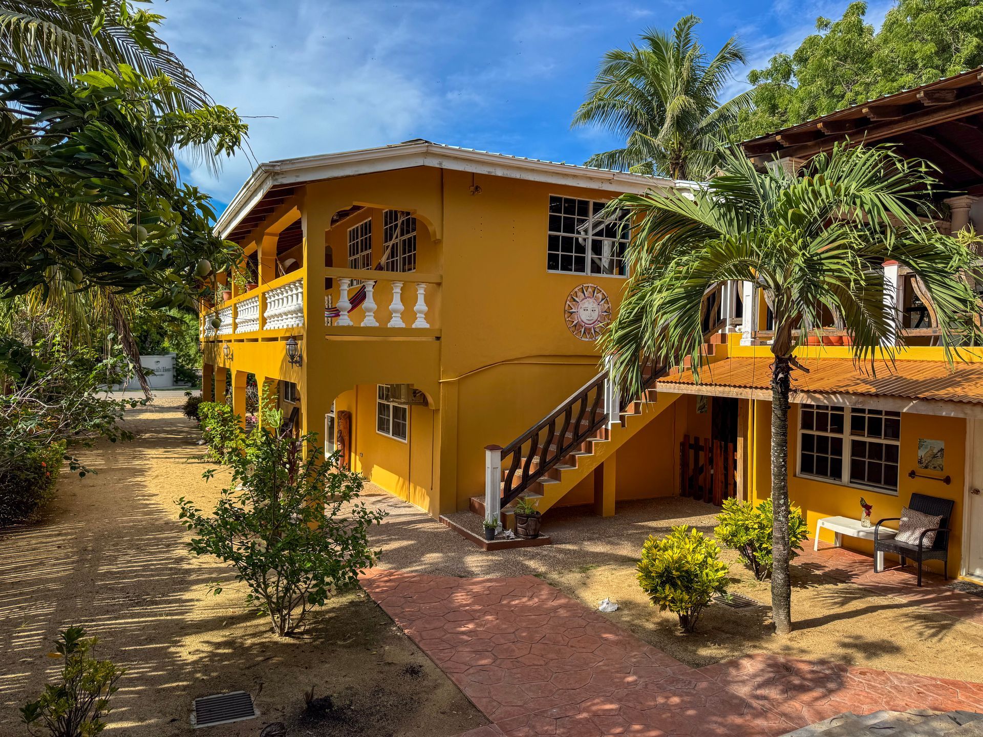 A large yellow house with stairs leading up to the second floor is surrounded by palm trees.