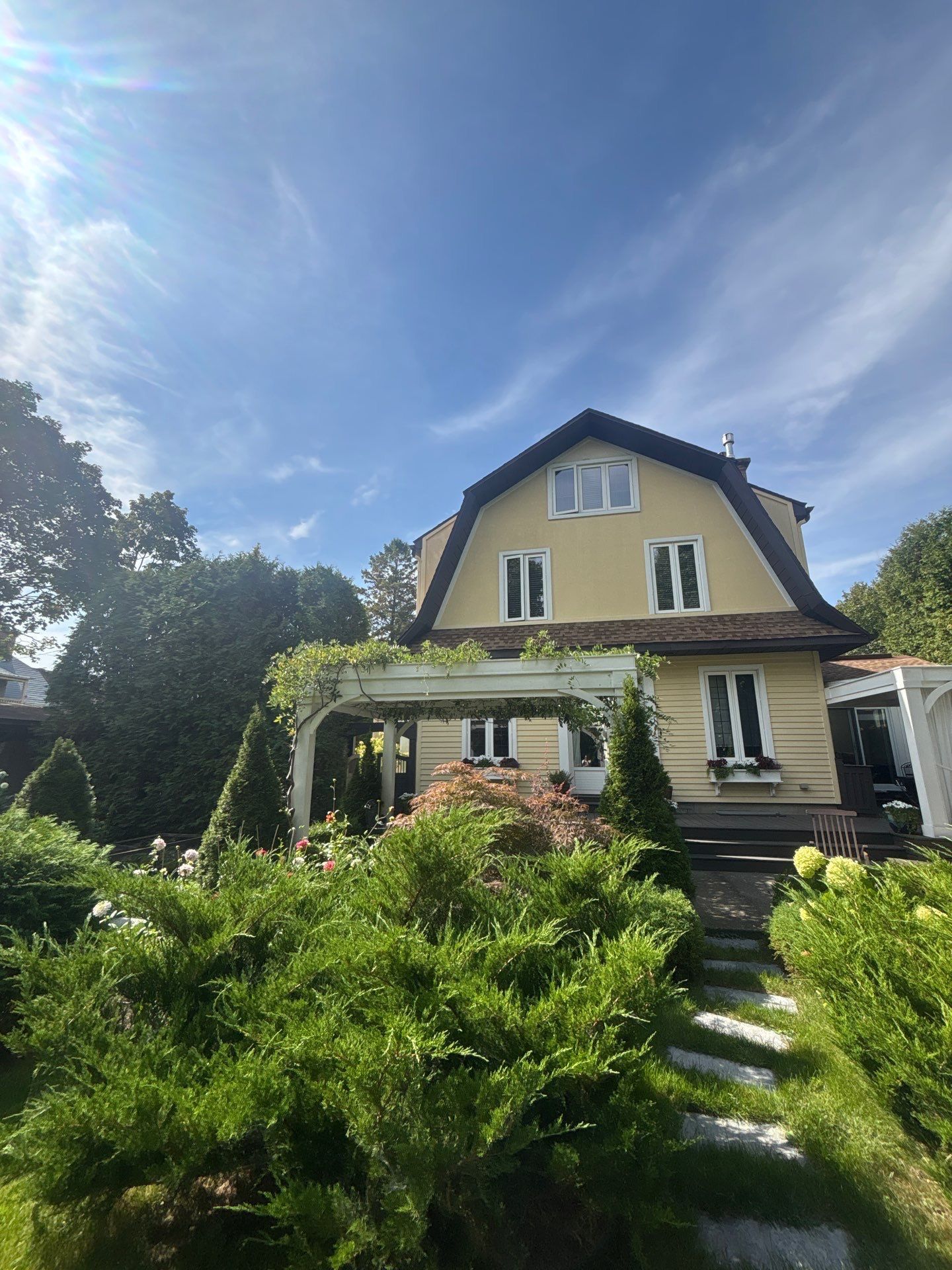 Maison jaune aux boiseries blanches et à la véranda, entourée d'une végétation luxuriante sous un ciel d'un bleu éclatant.