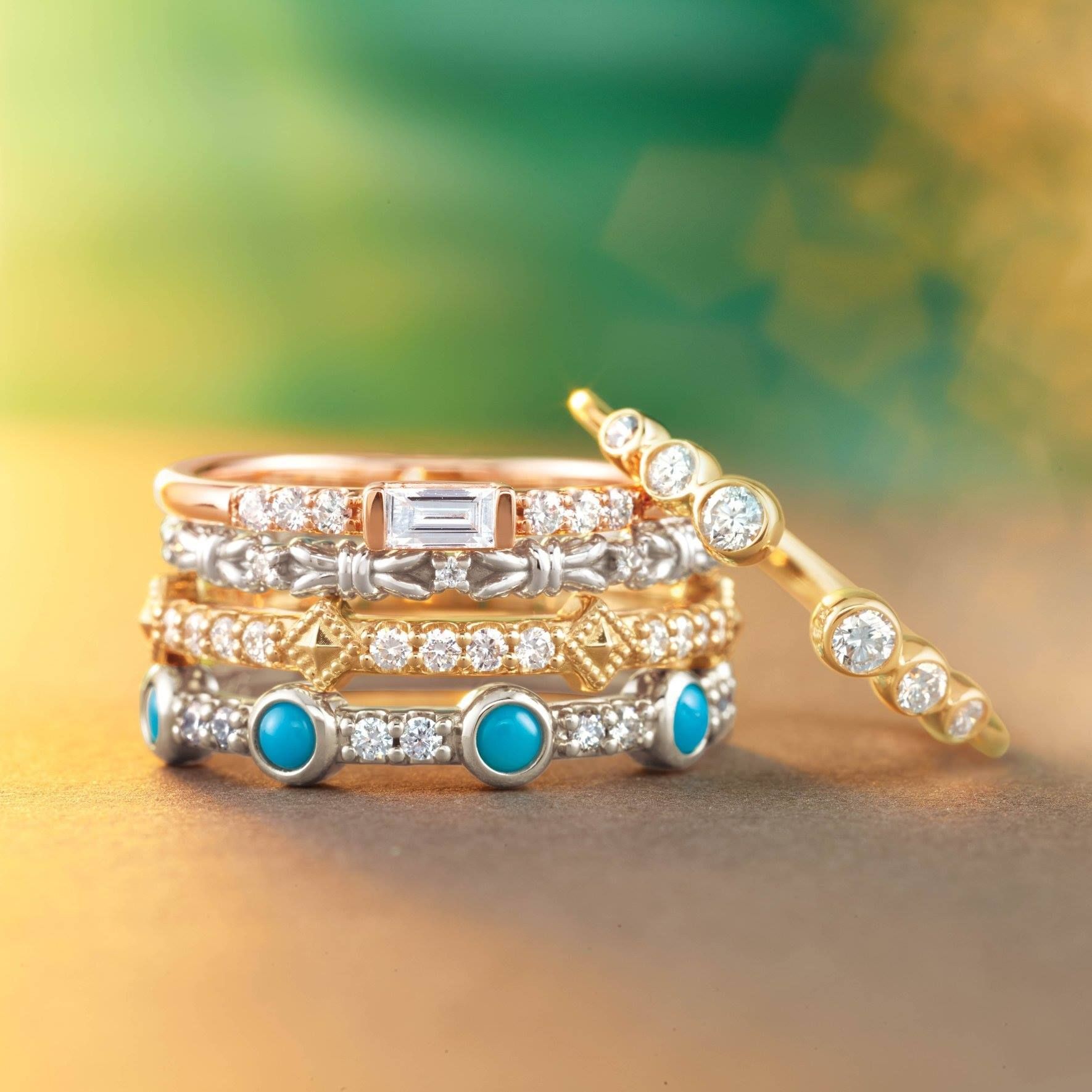 Stack of colorful gemstone and diamond rings on a warm, blurred background.