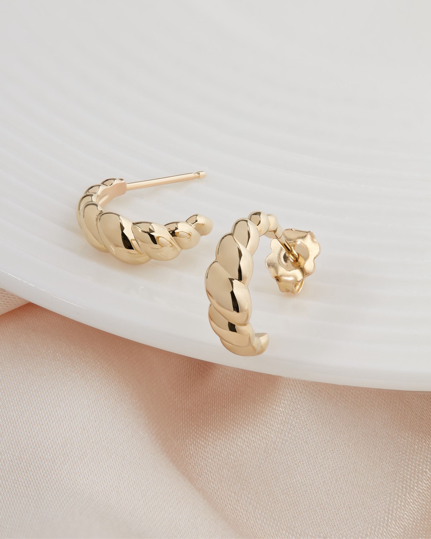 Gold croissant-shaped hoop earrings on a white surface with a pink cloth background.