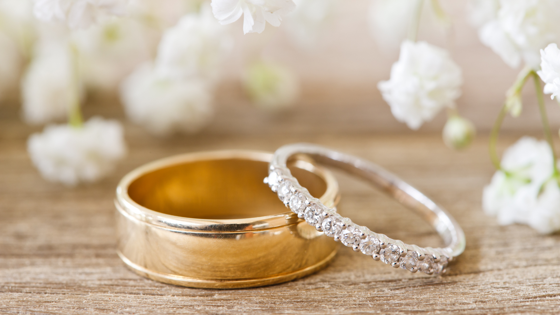 Two wedding rings, gold band and diamond band, on wood surface, with white flowers.