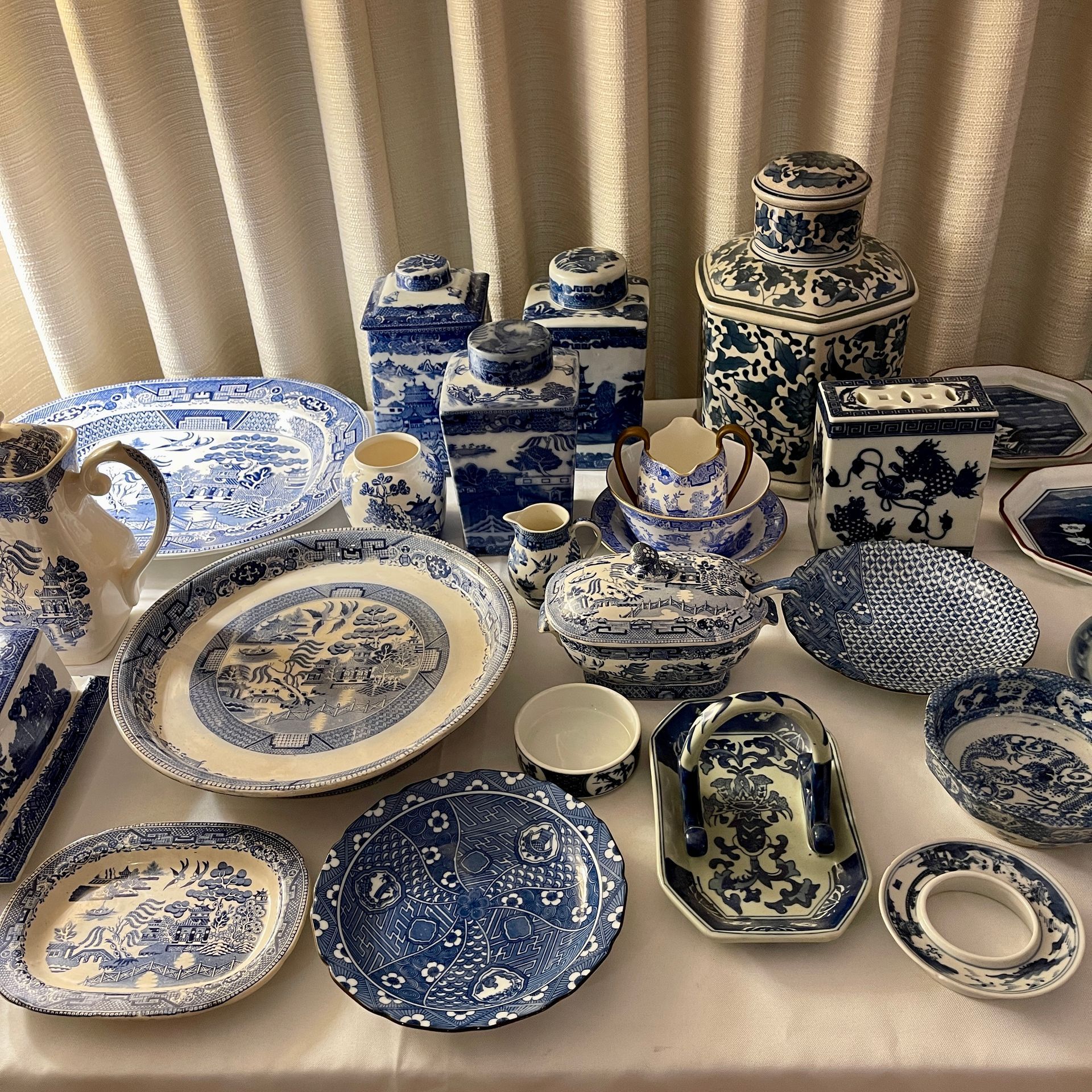 Blue and white porcelain dishes on a table, including platters, pitchers, and jars, arranged against a white curtain.