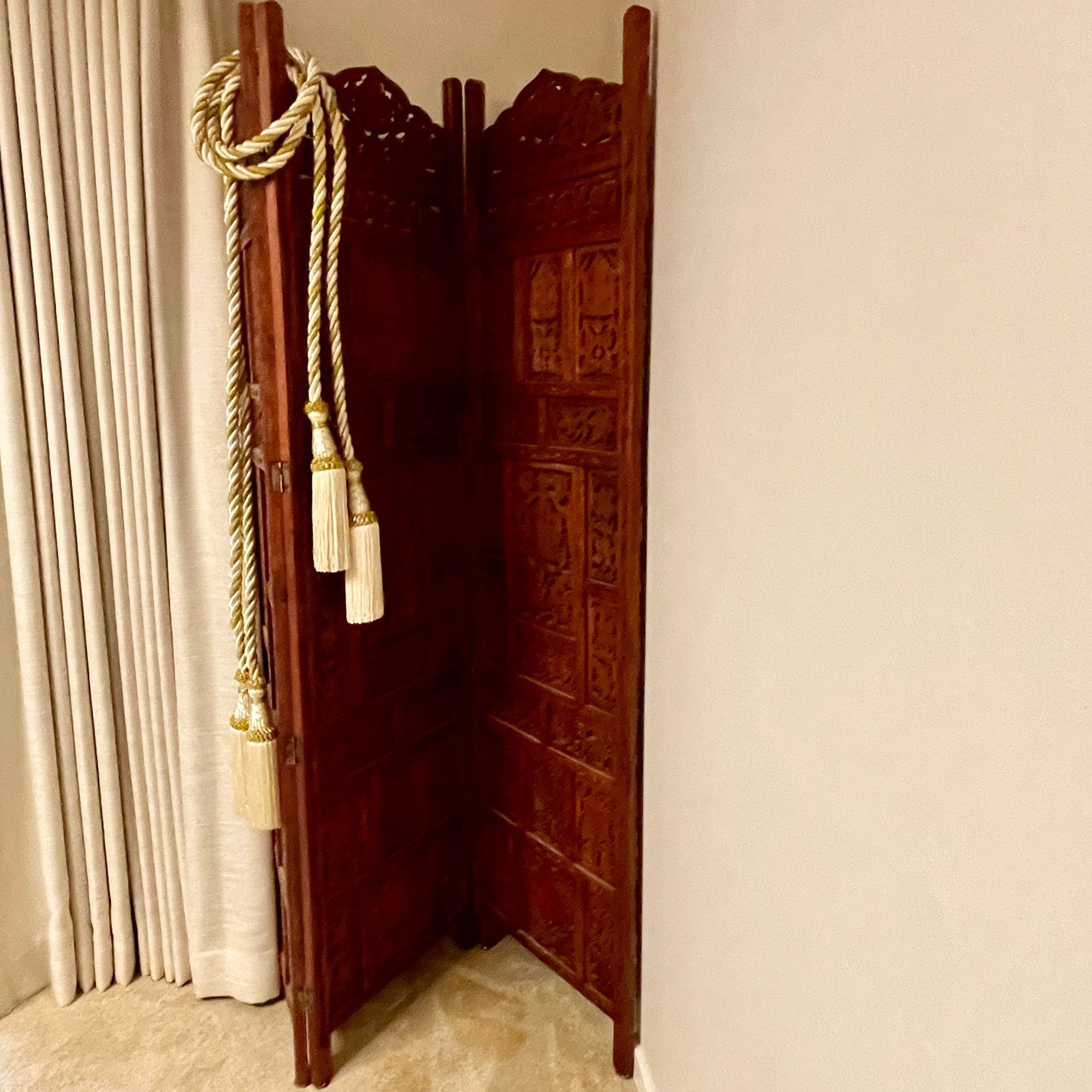 Dark wooden folding screen with carved details, next to a beige curtain. A gold rope with tassels hangs on it.
