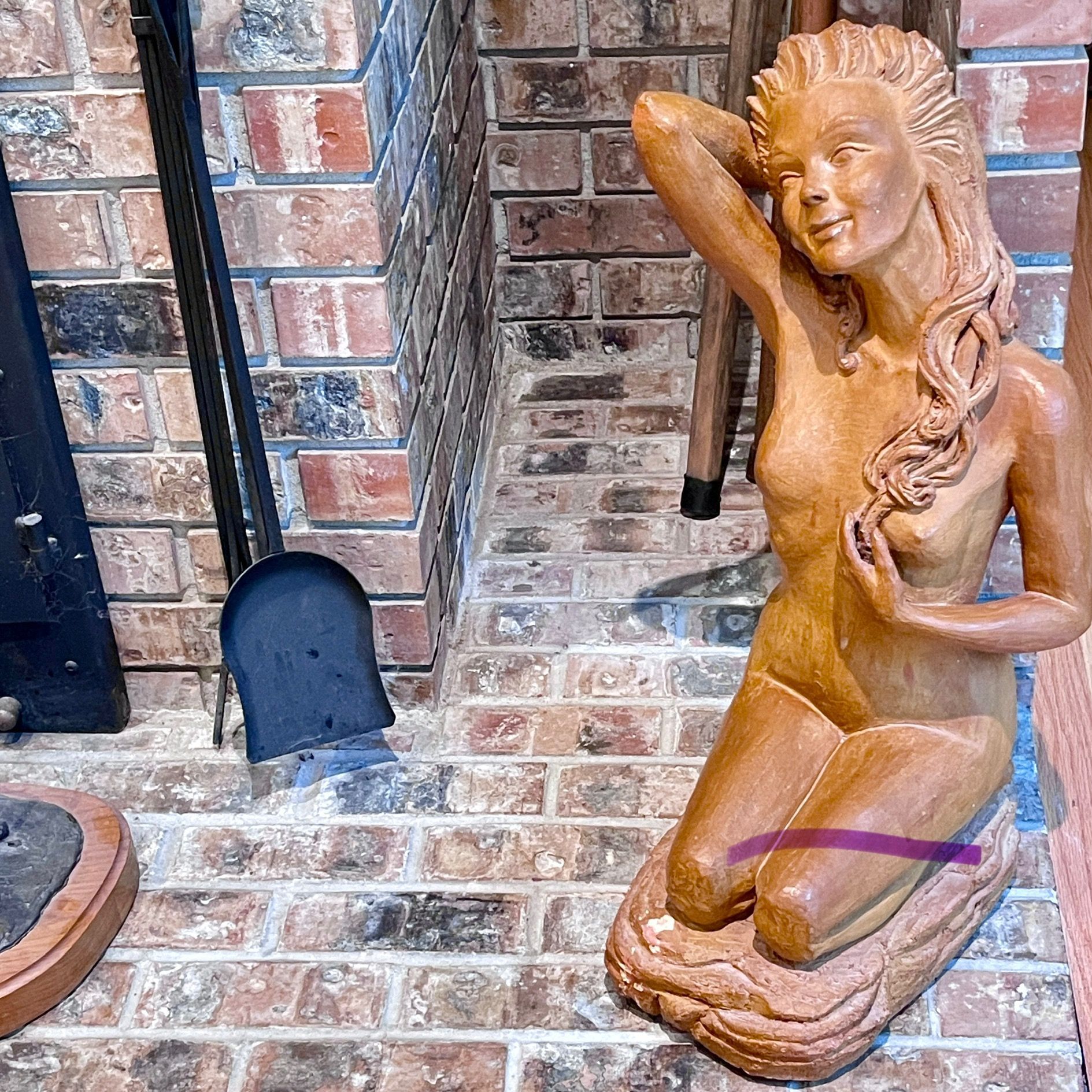 Wooden mermaid statue kneeling by a brick fireplace, smiling with arm raised.