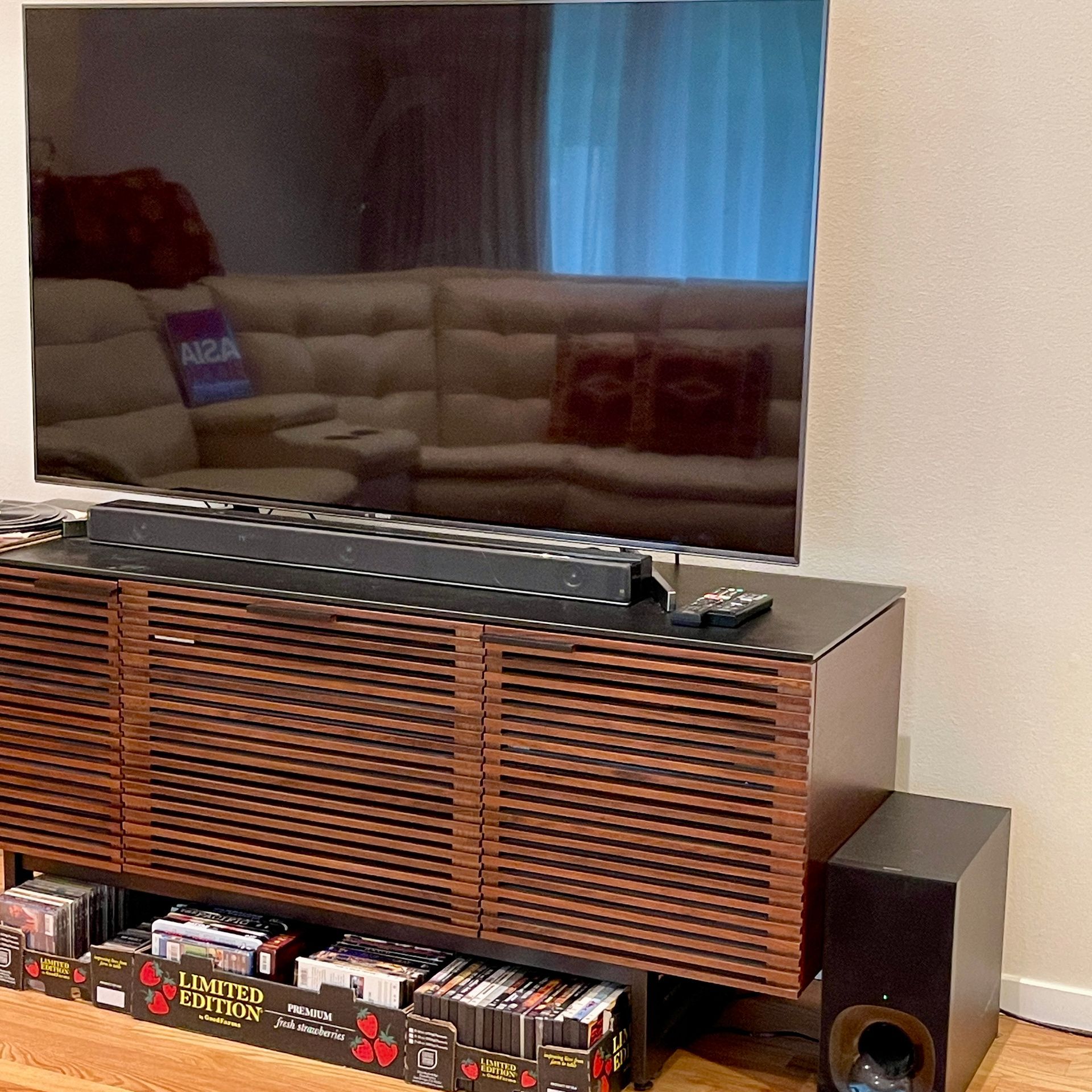 A large TV sits above a soundbar on a brown console. A subwoofer is to the right. A beige sectional is reflected in the TV.