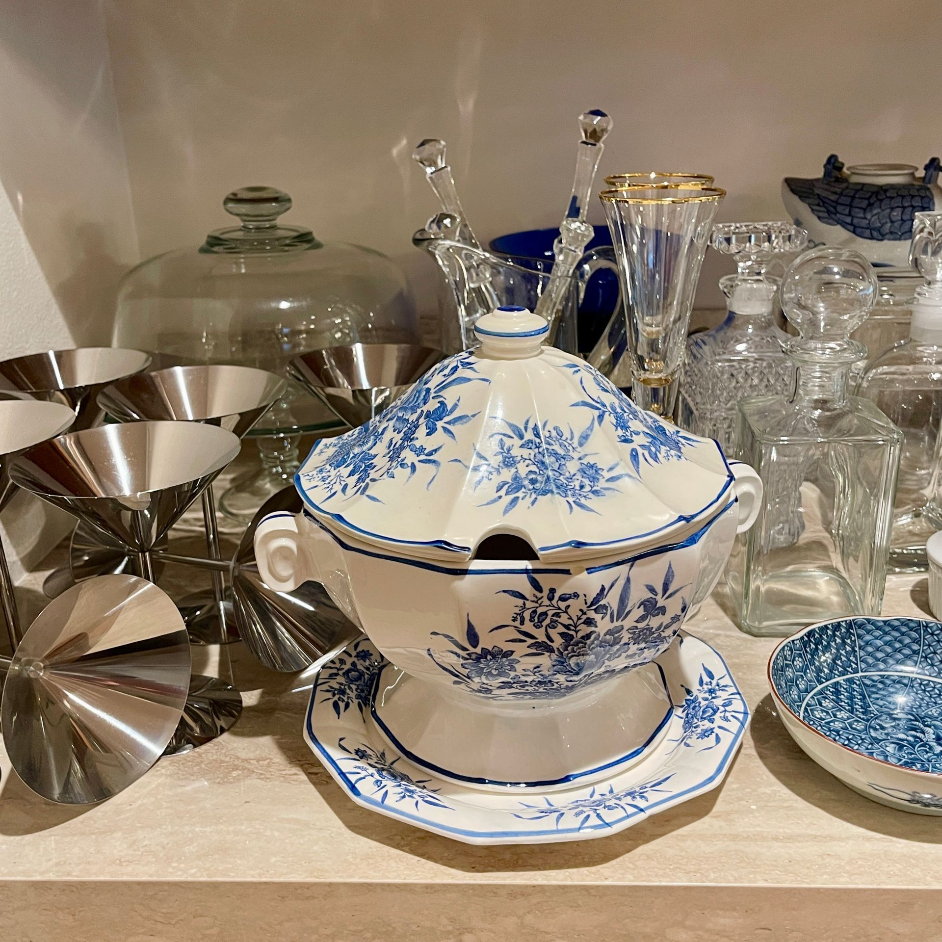 Blue and white floral soup tureen on a shelf with glass and metal serving items.