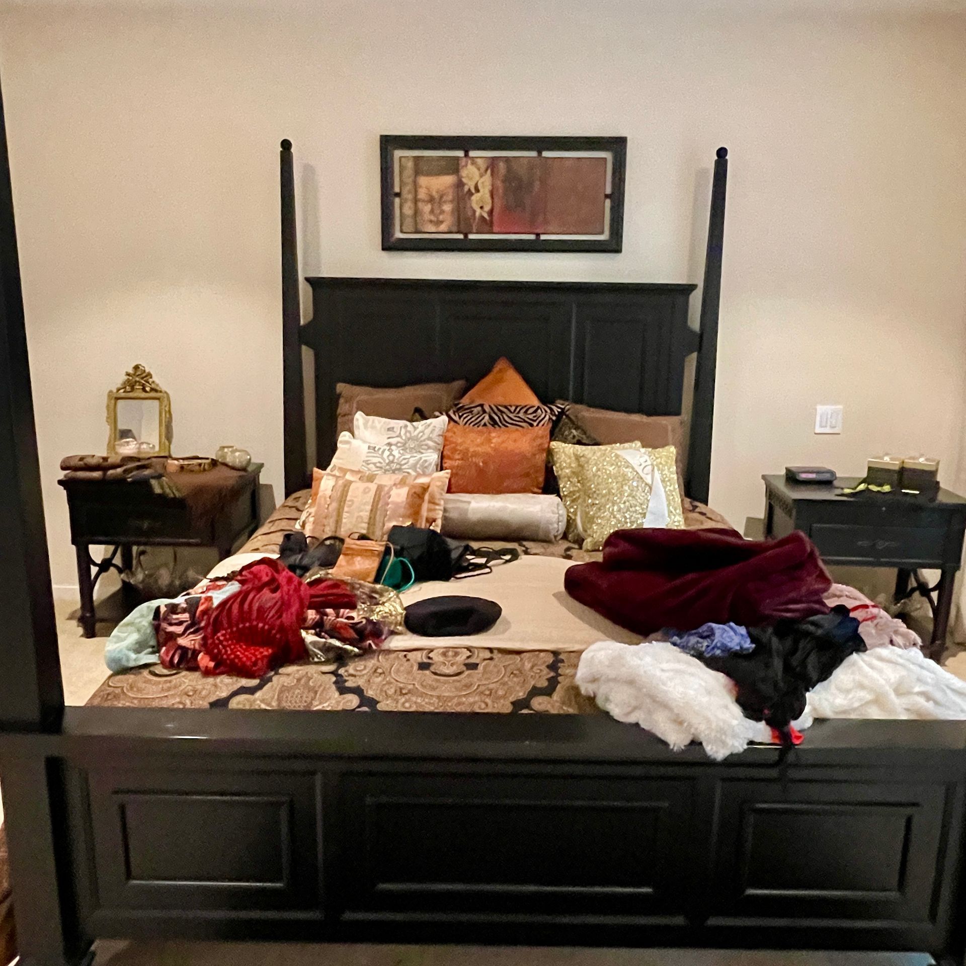 Black four-poster bed with patterned rug, pillows, and scattered clothing. Two nightstands flank the bed.