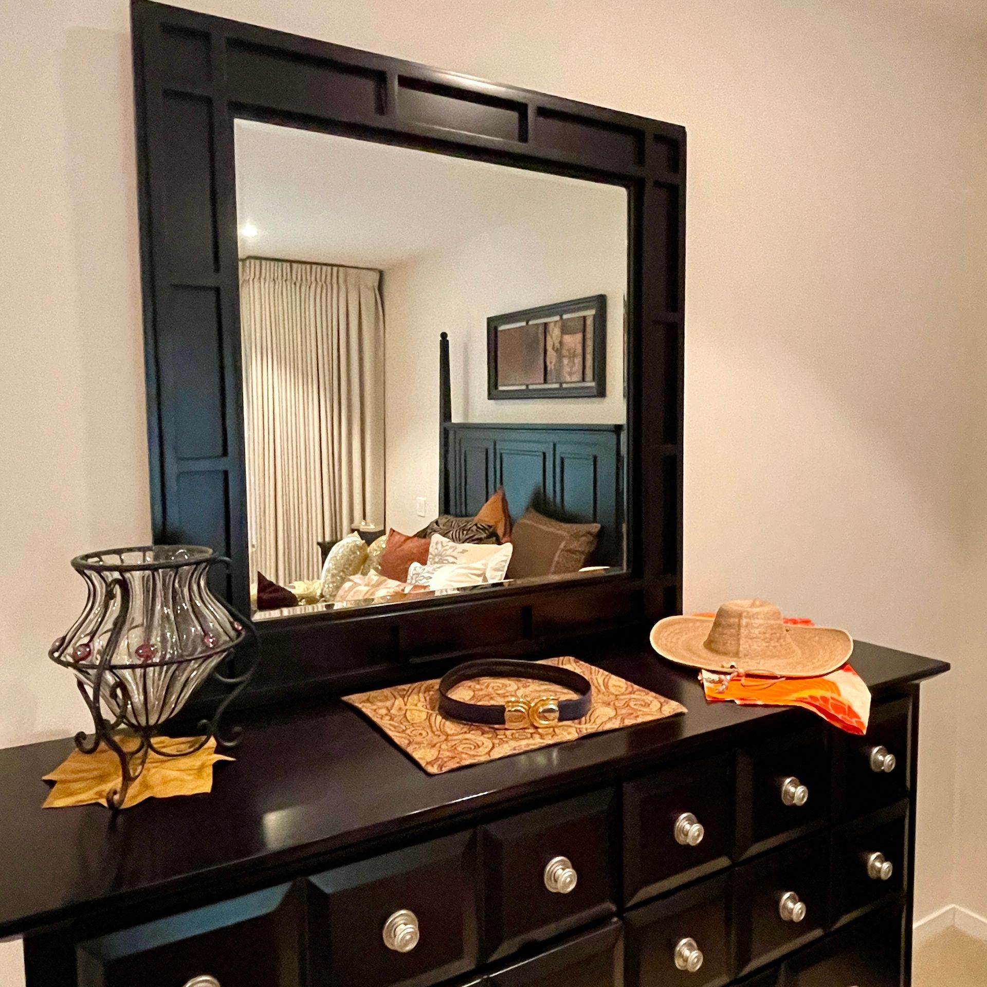 Black dresser with mirror reflecting bedroom with bed and curtains. A hat and belt sit on the dresser.