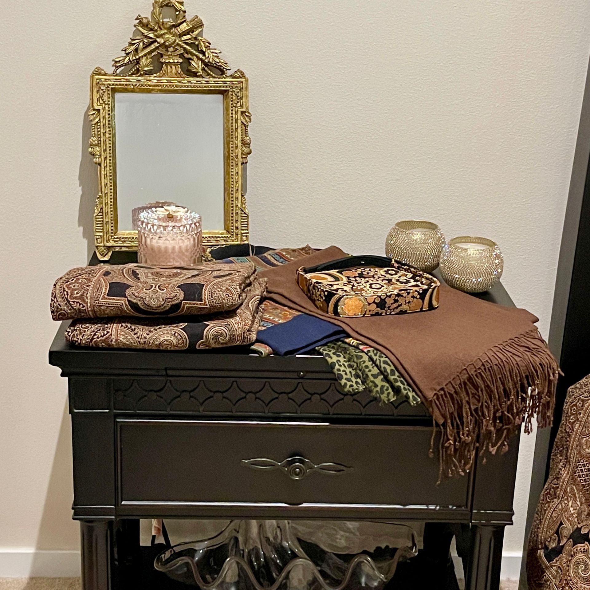 Nightstand with a gold mirror, folded fabrics, candles, and a leopard print box.