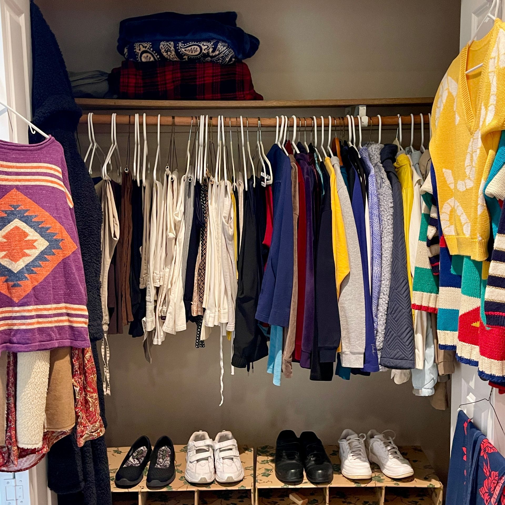 Clothes hanging in a closet with shoes below, stacked clothes above, and a colorful knit sweater on the right.