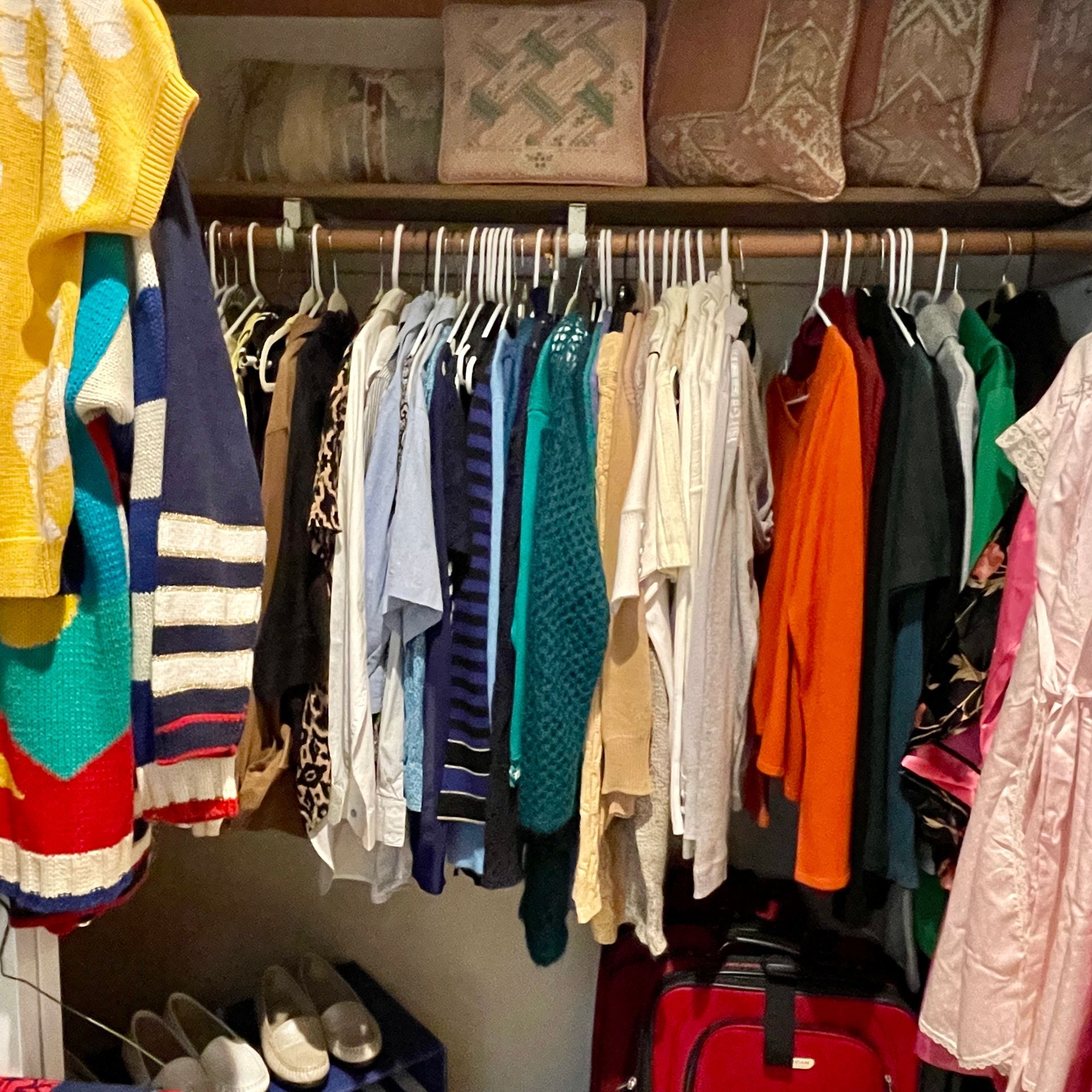 Closet filled with various clothing items on hangers; luggage and shoes below, pillows above.