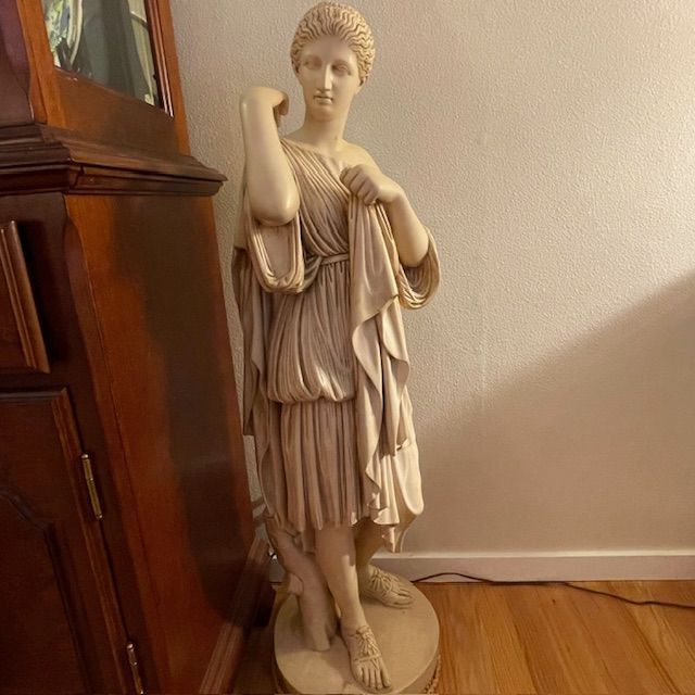 Statue of a draped classical figure, cream-colored, standing on a circular base.