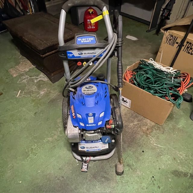 Blue Subaru pressure washer with gray handle and attached cords; next to a box with green cords.