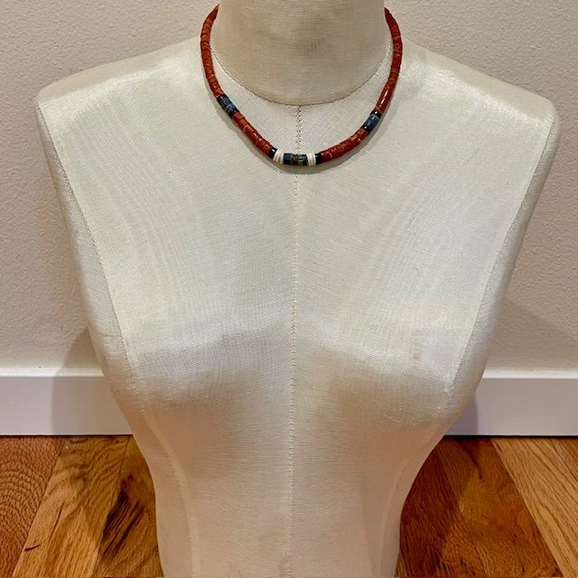 A red braided necklace on a white dress form. It has a central white, black, and blue beaded accent.
