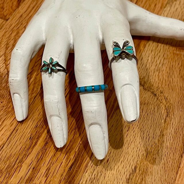 White hand statue with three rings: turquoise flower, band, and butterfly, on a wood surface.
