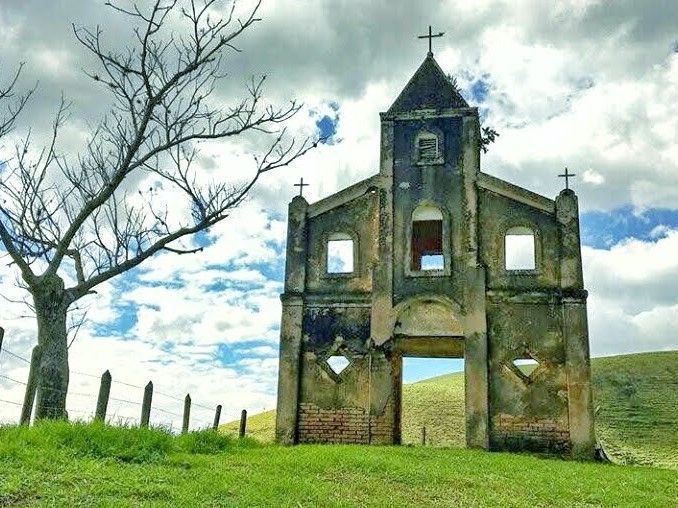 Fachada de igreja de pedra em ruínas numa colina gramada sob um céu nublado, com uma árvore nua e postes de cerca nas proximidades.