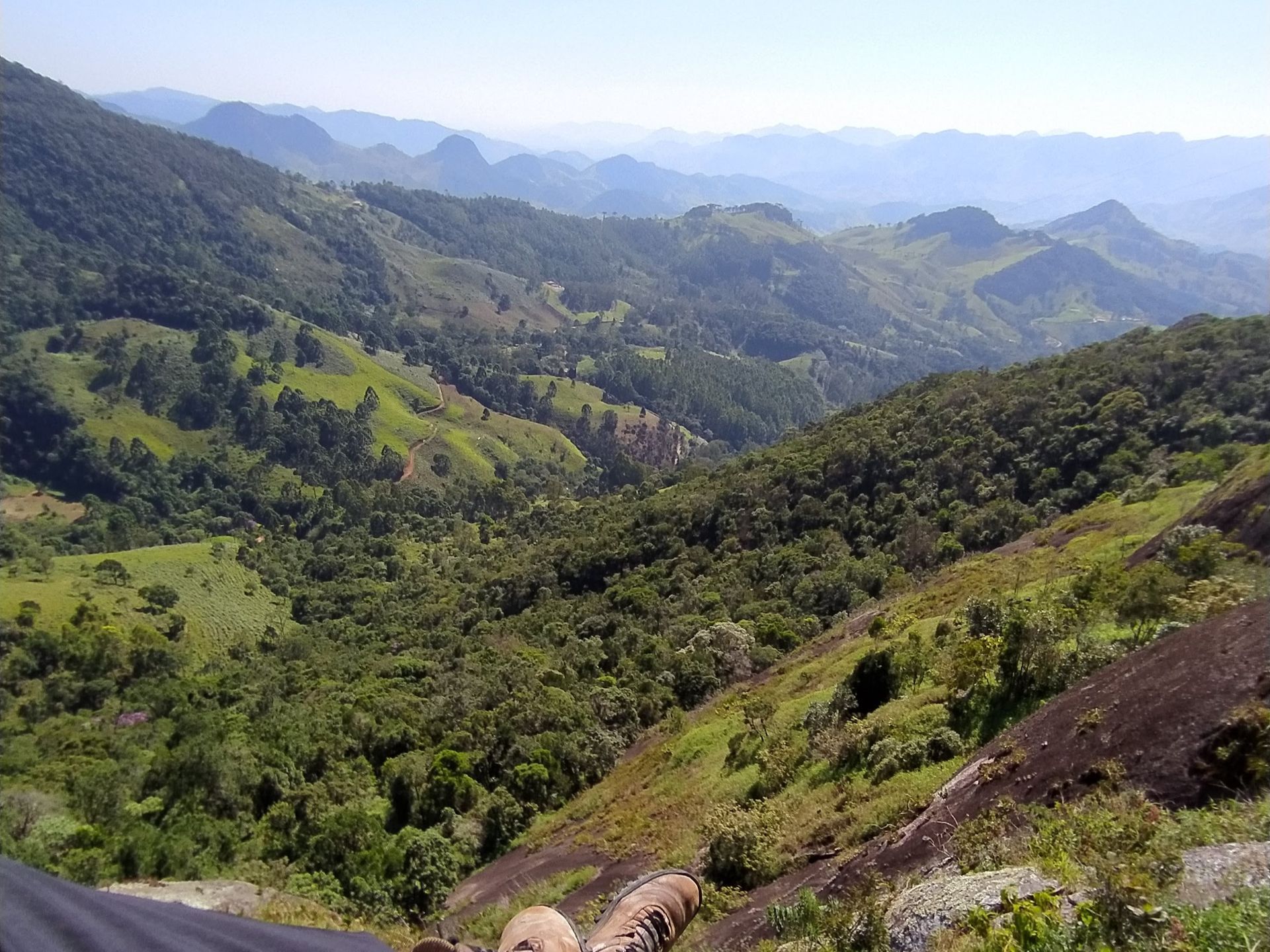 Vale verdejante das montanhas com colinas em camadas, encostas florestadas e cristas azuladas e nebulosas sob um céu brilhante.