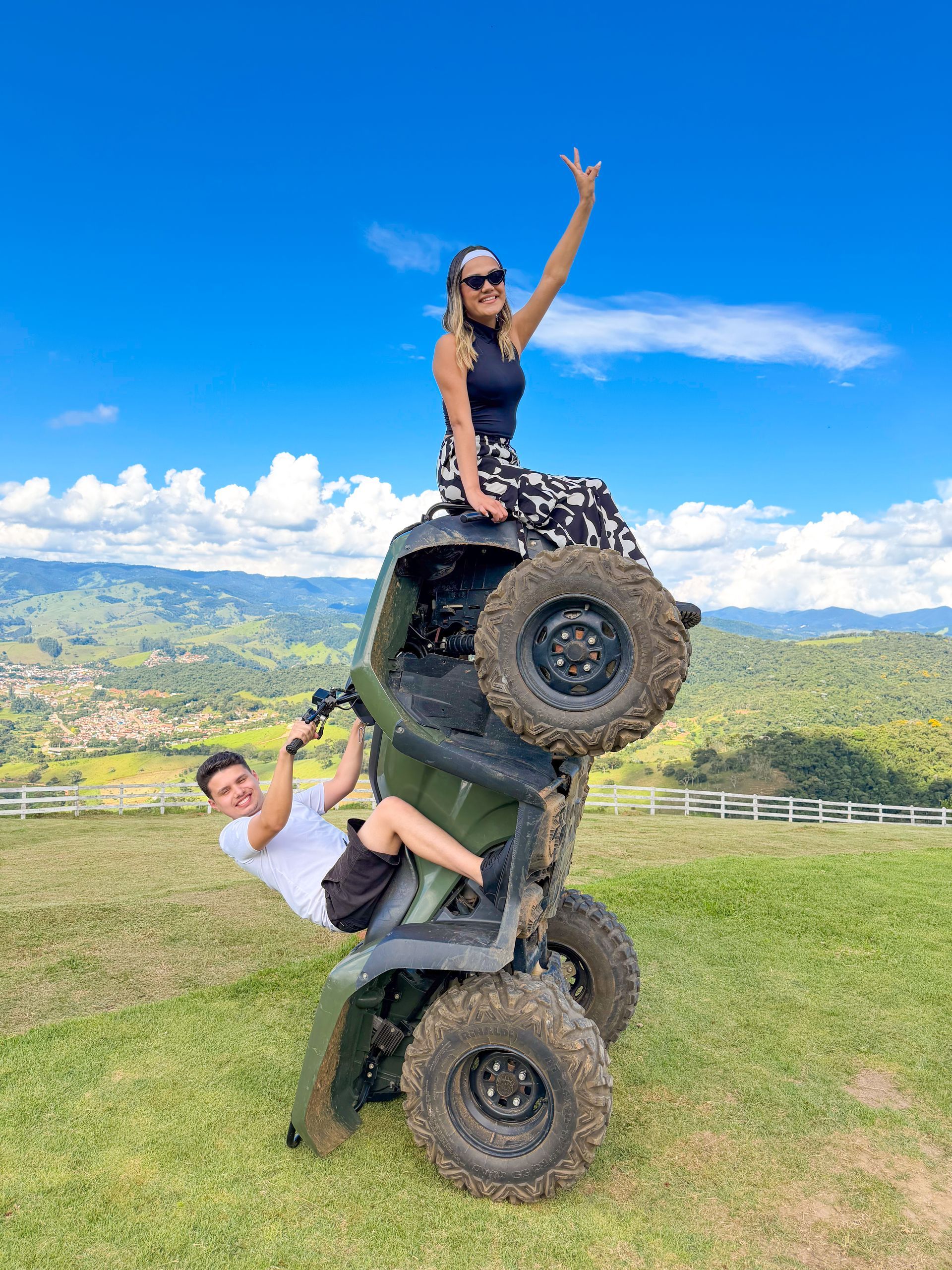 Duas pessoas posando em uma escultura feita com pneus de caminhão empilhados, em uma paisagem montanhosa gramada sob um céu azul.