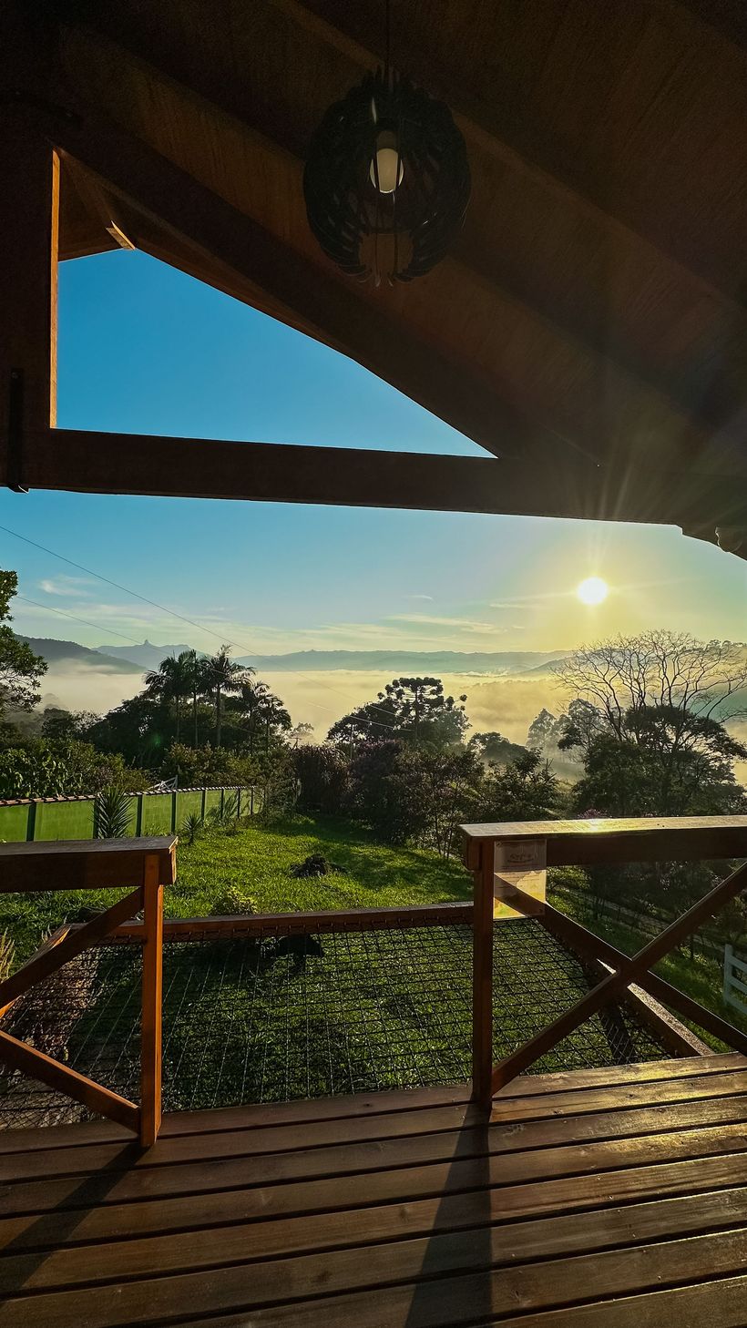 Deck de madeira iluminado pelo sol com vista para colinas e árvores envoltas em névoa ao nascer do sol.