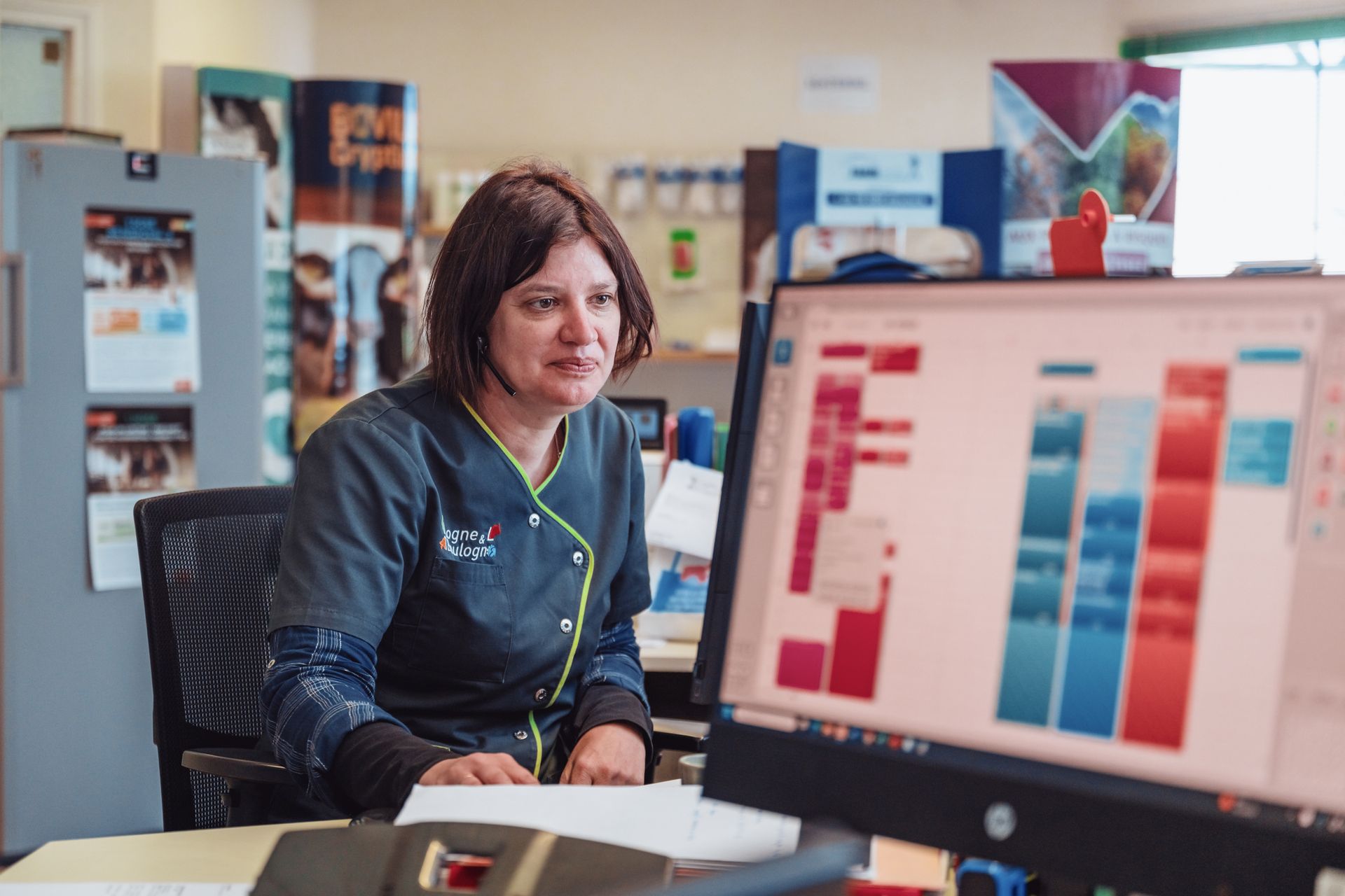 Une femme en blouse médicale regarde un écran d'ordinateur, probablement en train de planifier. Ambiance de bureau avec des objets colorés visibles.