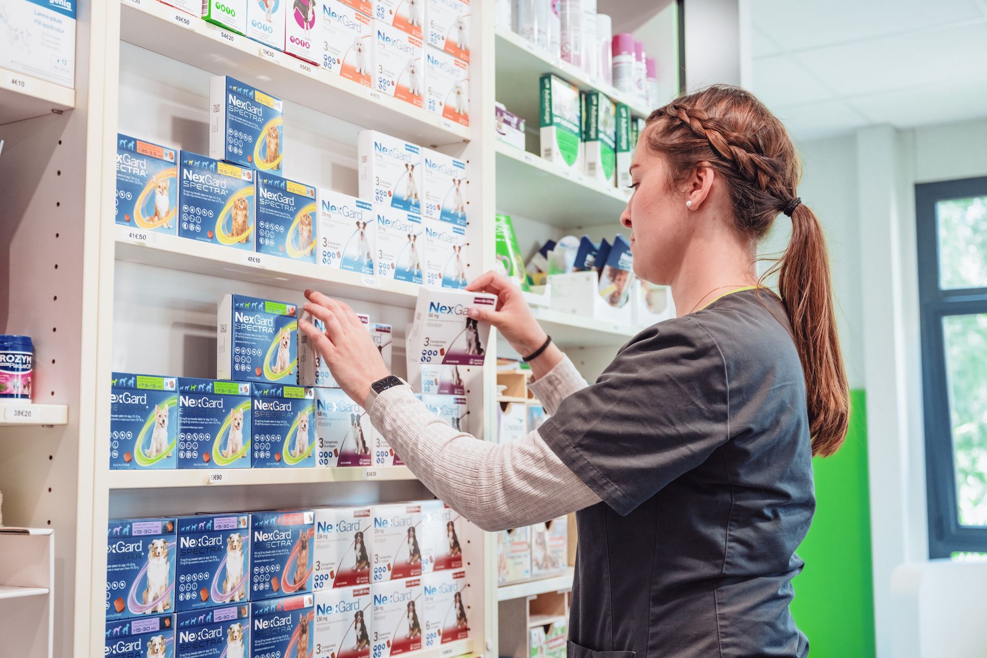 Une ASV remplit les rayons d'une pharmacie vétérinaire et prend un produit. Les rayons sont remplis de divers articles emballés.