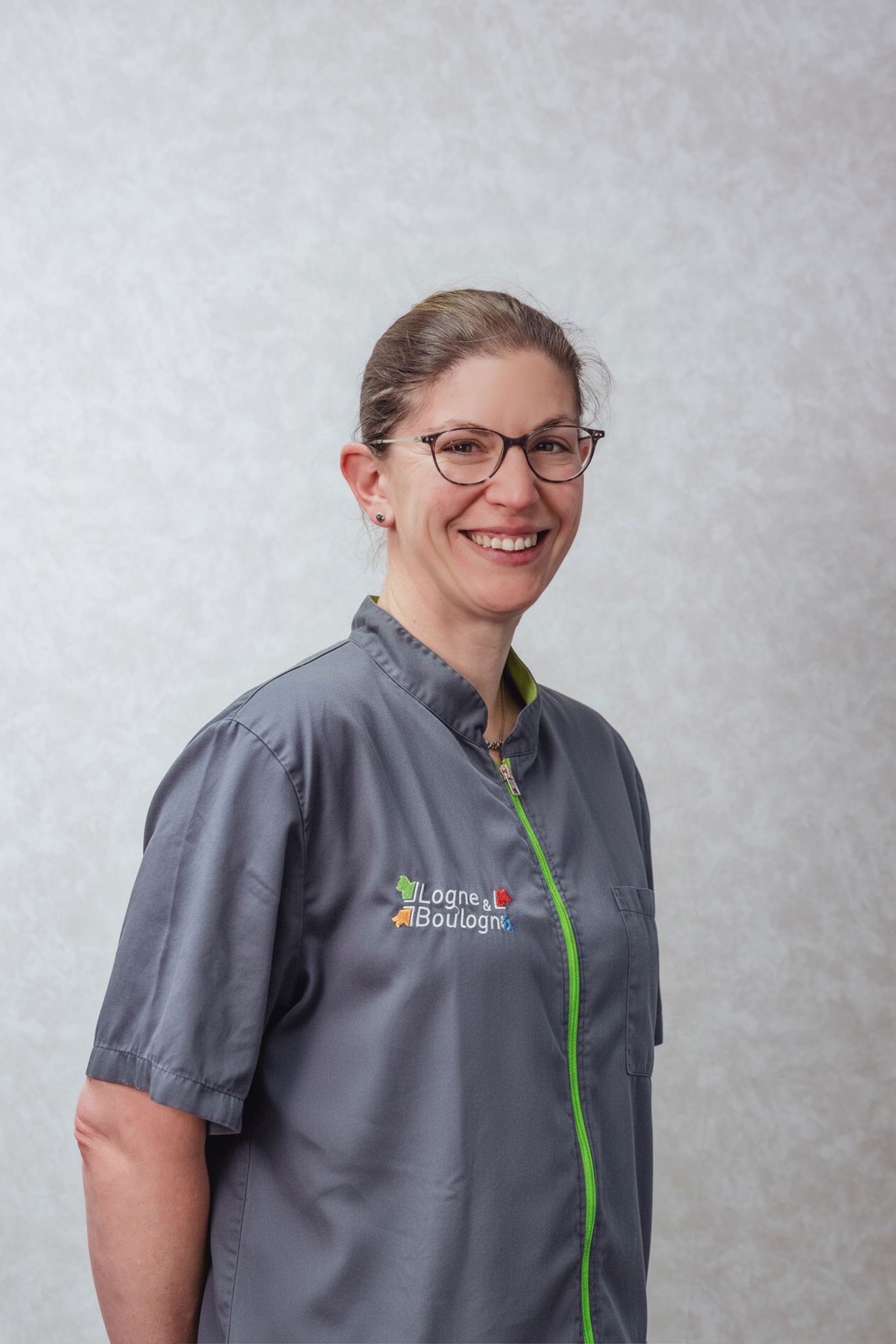 Femme en uniforme gris souriante, portant des lunettes. Fermeture éclair et logo verts sur la chemise.