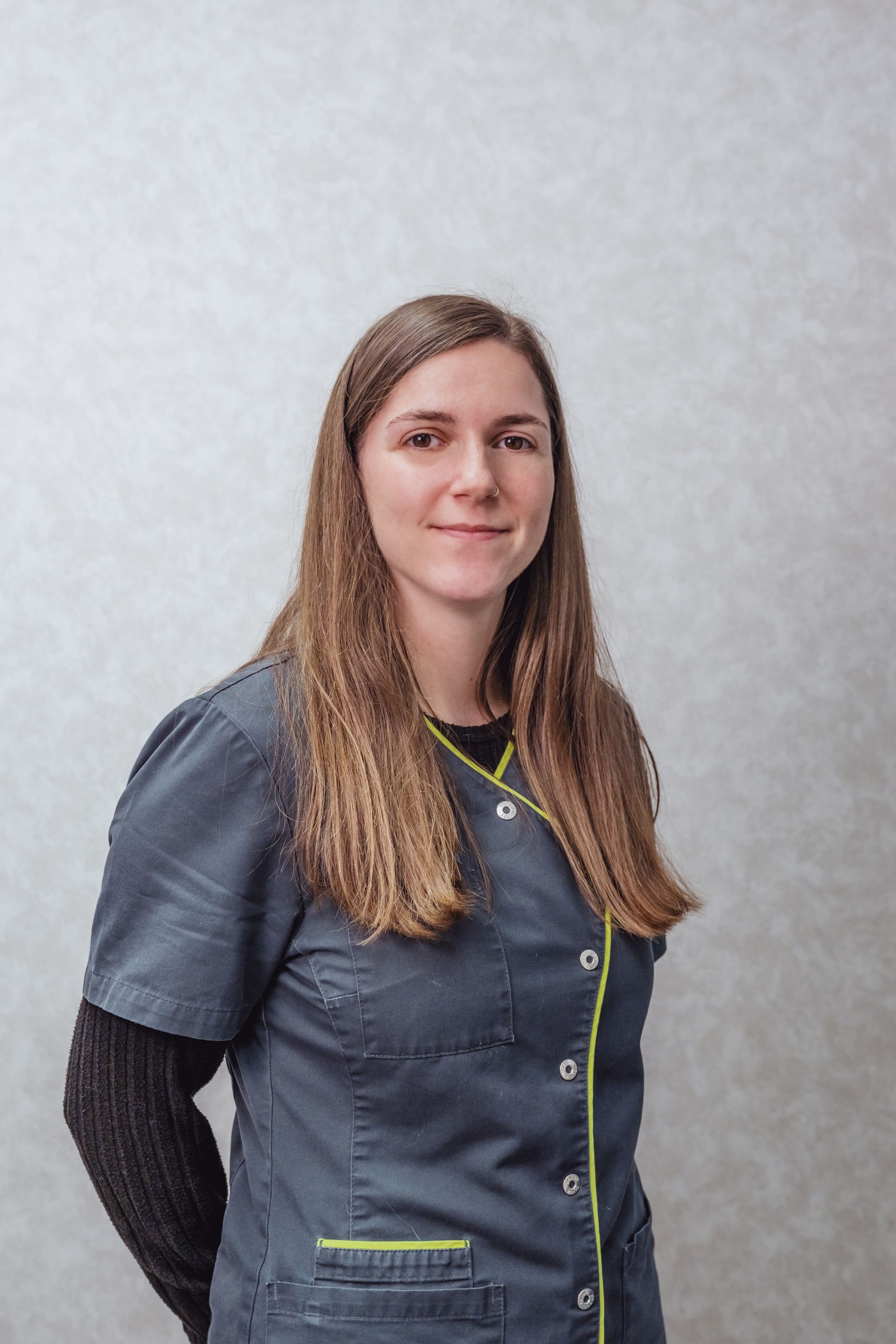 Femme en blouse bleue souriante, debout devant un fond neutre.