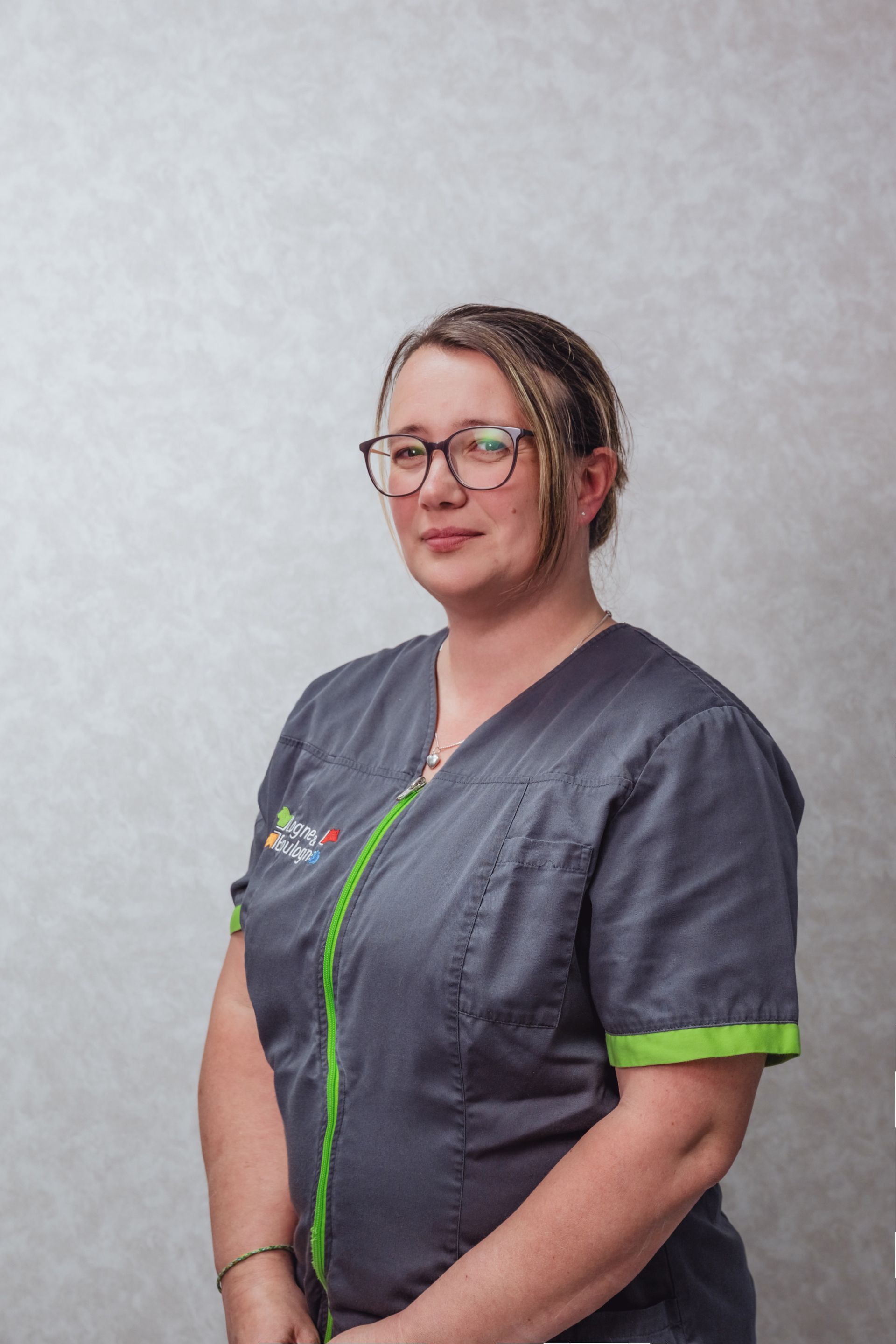 Femme en blouse grise à bordures vertes, portant des lunettes, esquissant un sourire.
