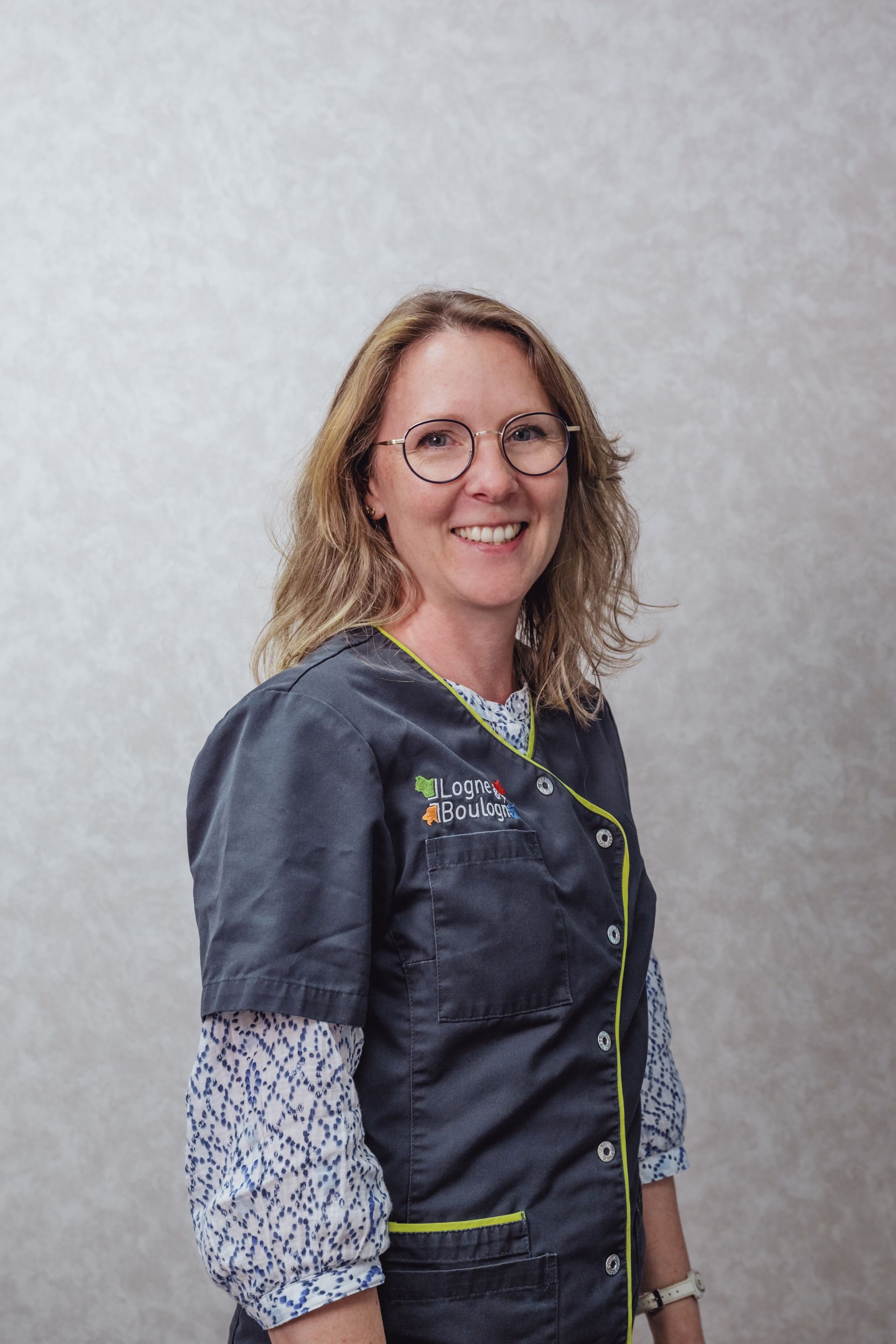 Une femme en uniforme bleu sourit, portant des lunettes, adossée à un mur à motifs.