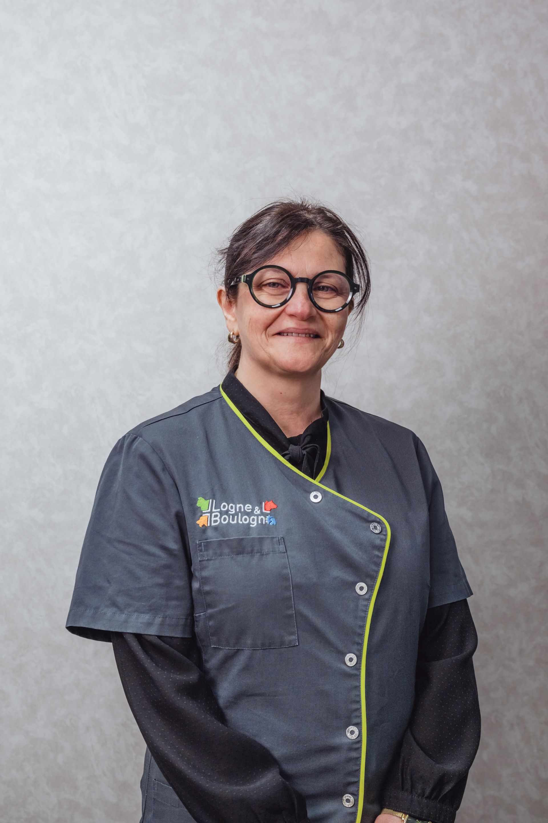 Une femme en uniforme bleu sourit, portant des lunettes, debout devant un mur de couleur neutre.