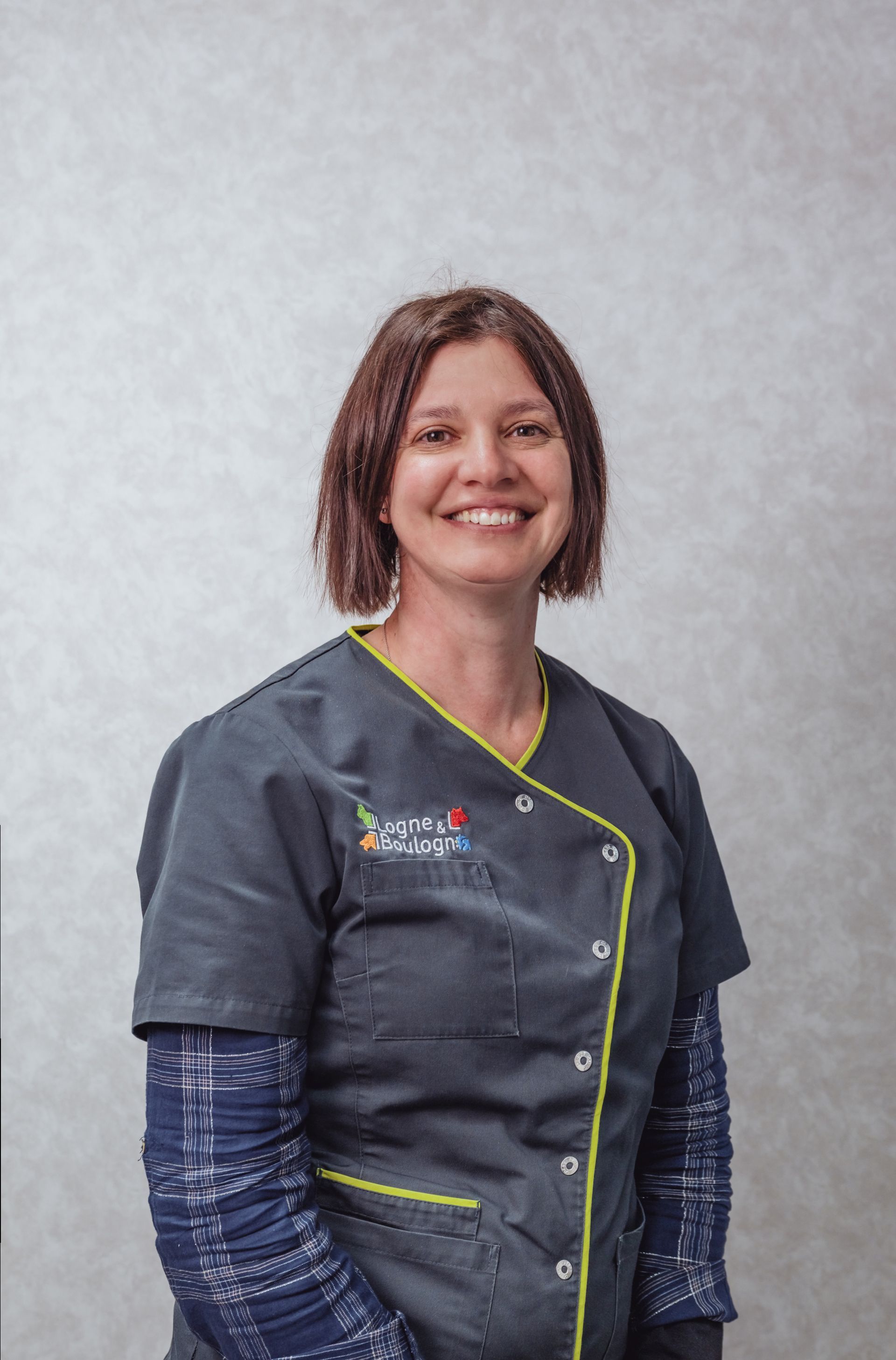 Une femme en blouse bleue sourit, debout à l'intérieur.