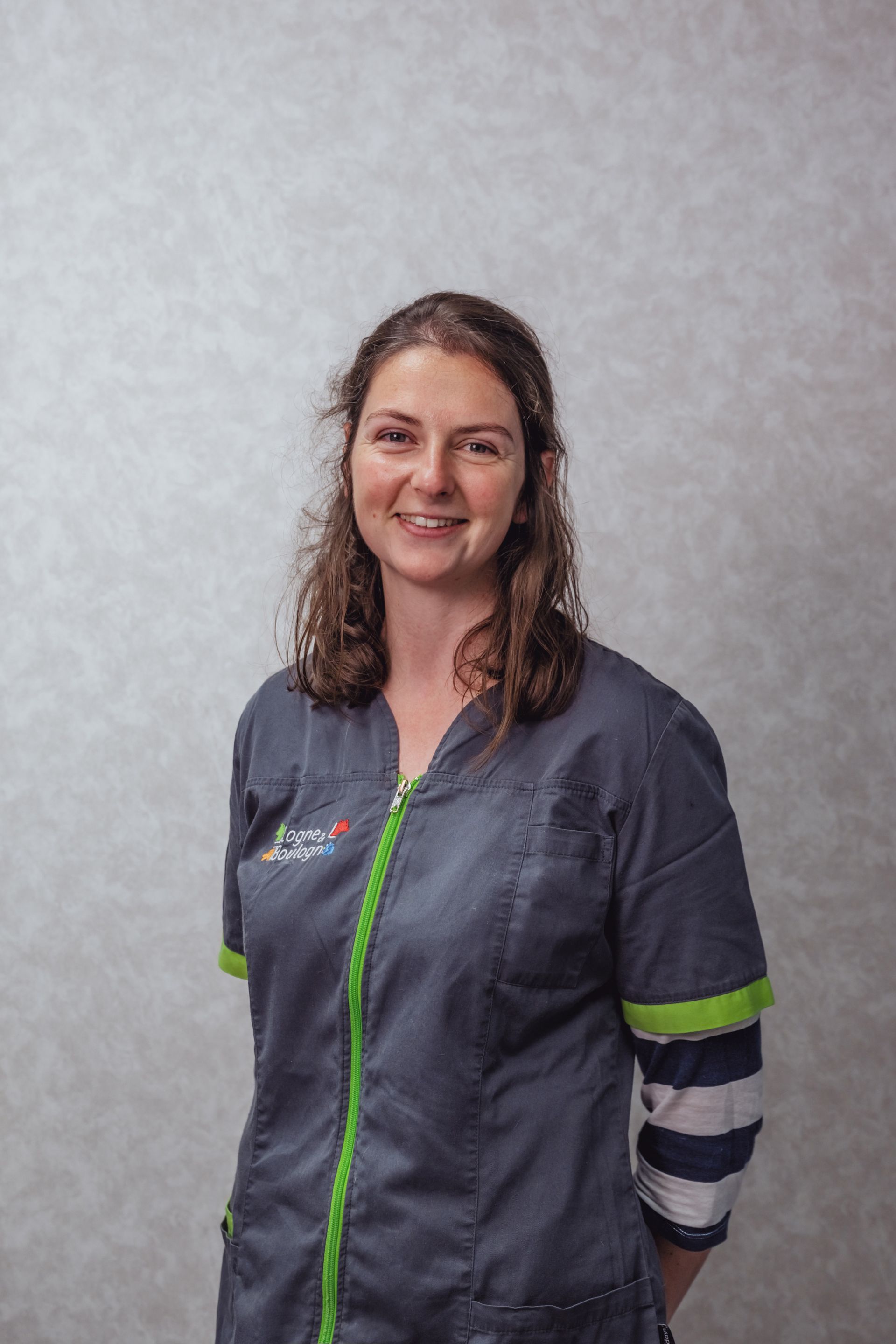 Une femme en uniforme gris sourit, adossée à un mur neutre.