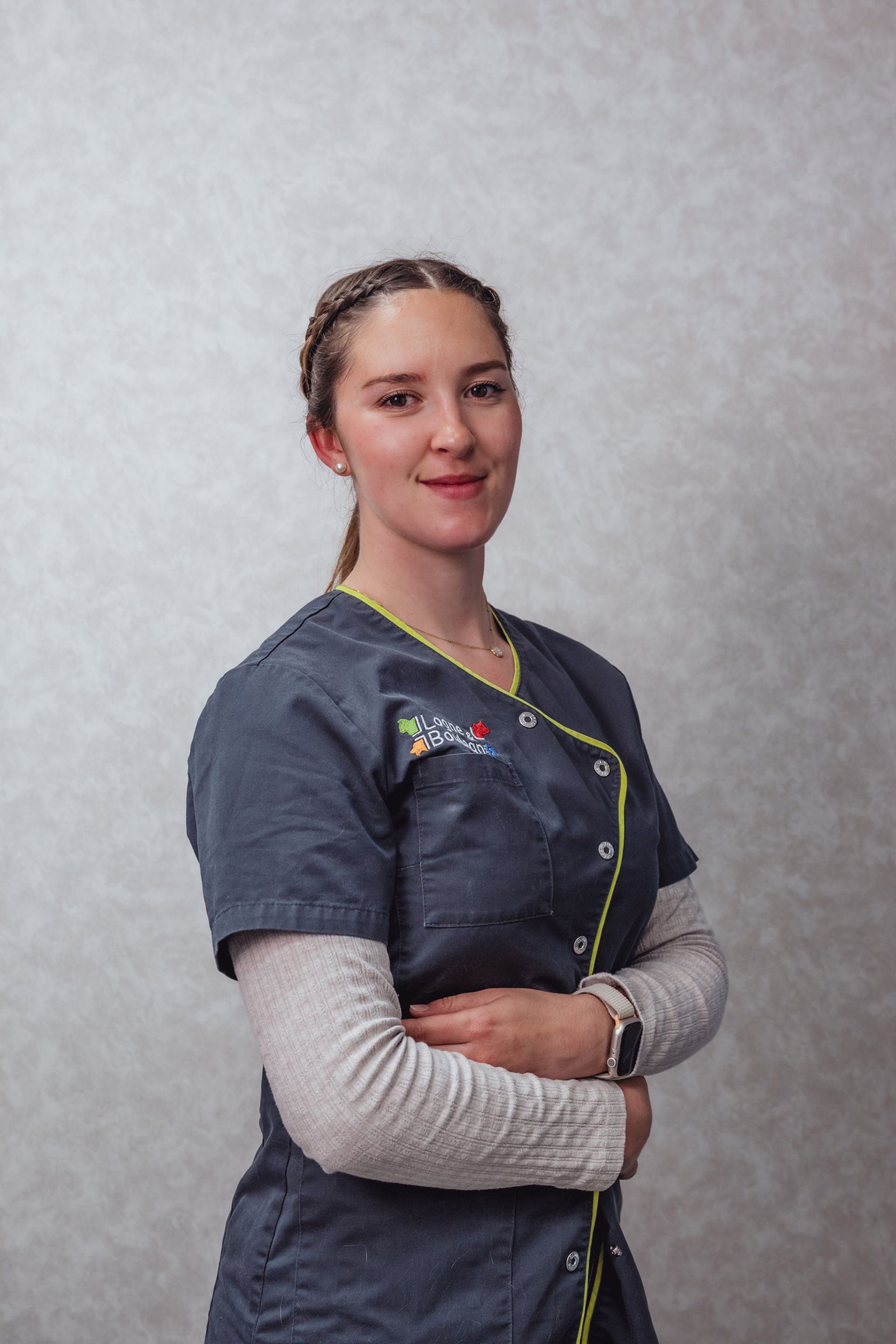 Femme en blouse médicale, les bras croisés, souriante. Cheveux clairs tressés.