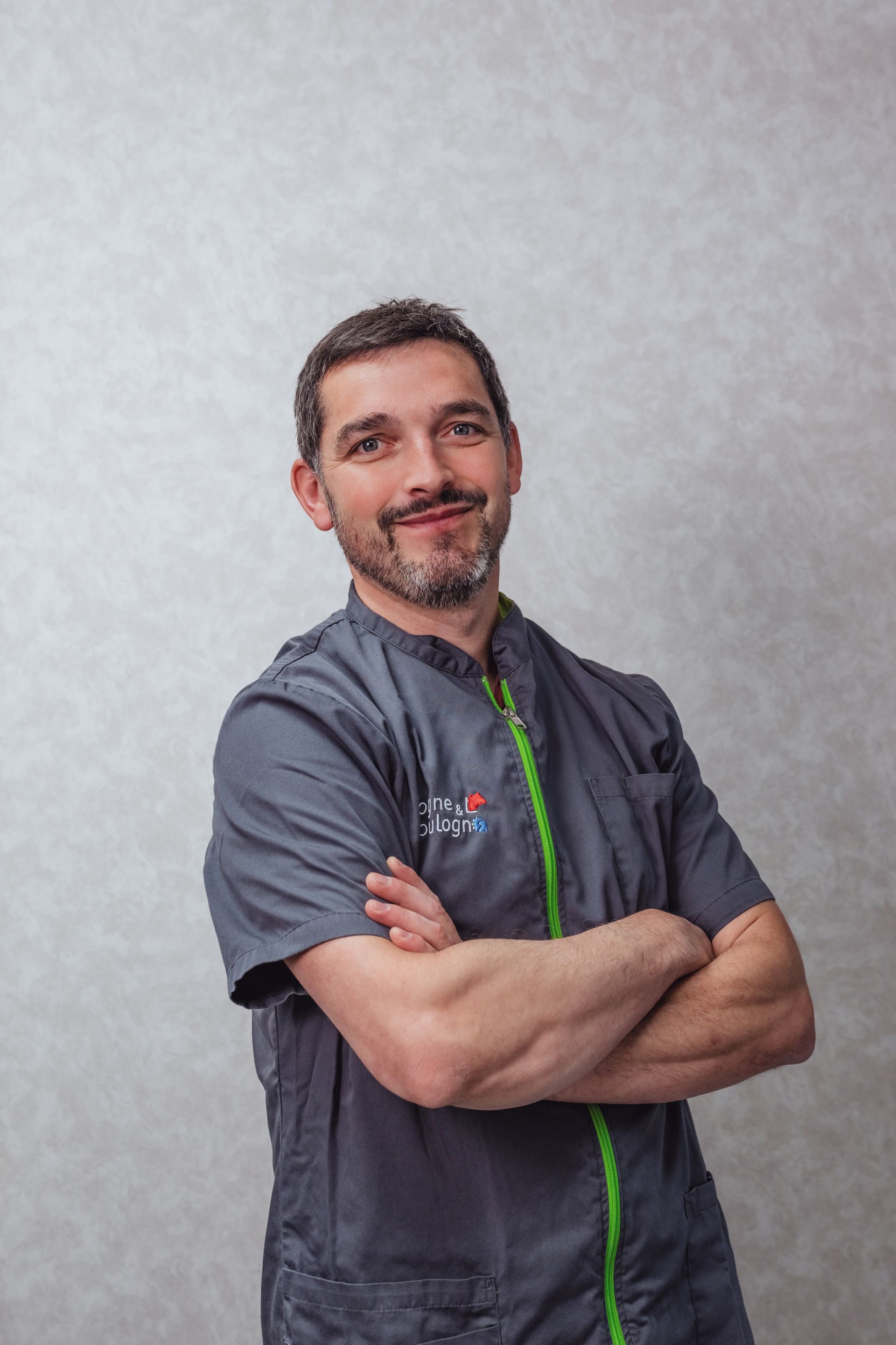 Un homme en uniforme gris, les bras croisés, souriant, se tient devant un mur gris clair.
