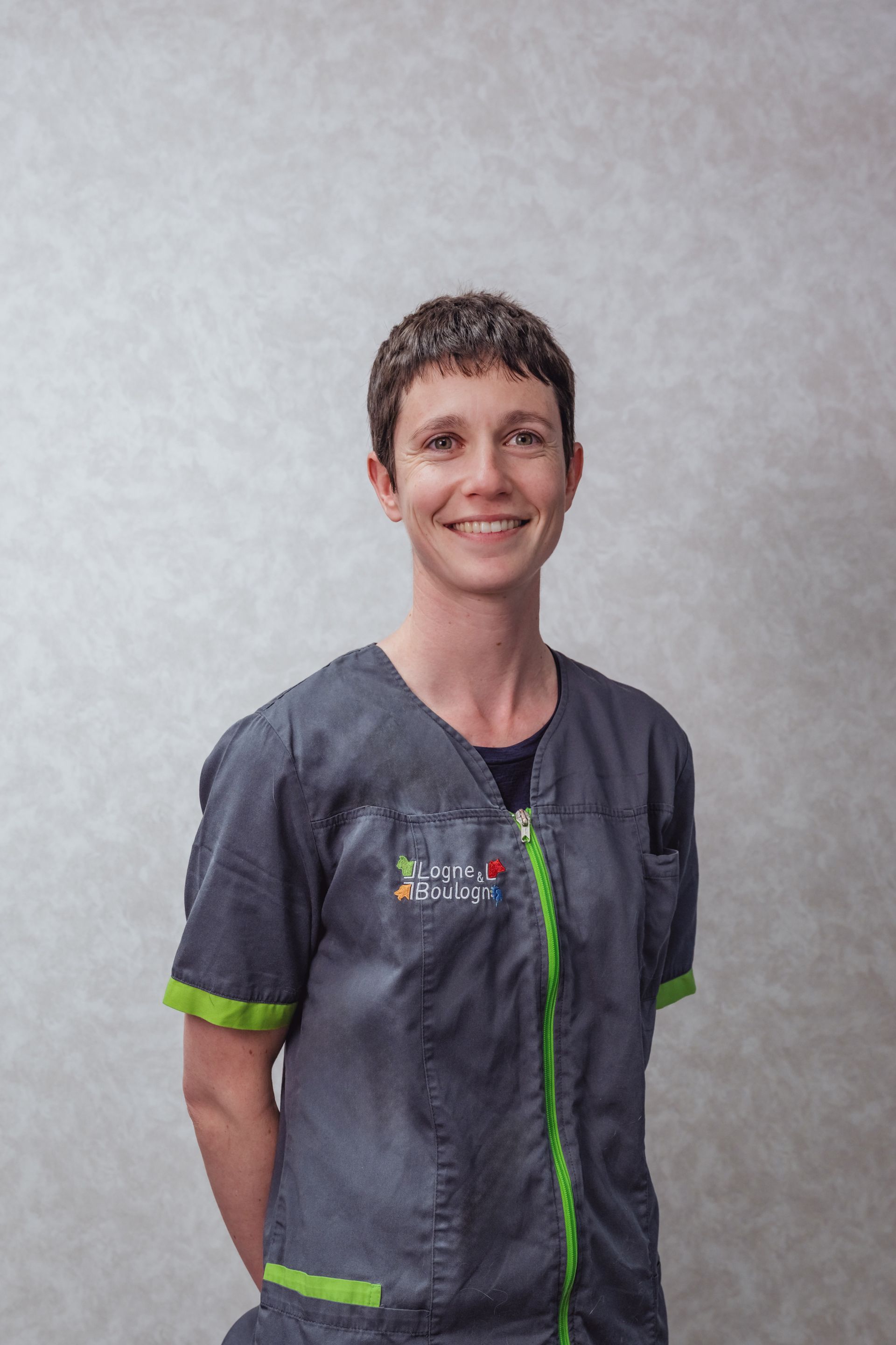 Une femme en blouse grise sourit, debout devant un mur texturé.