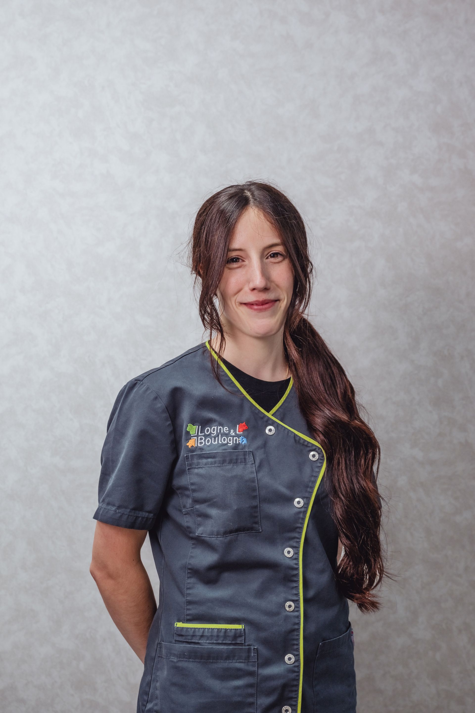 Femme en blouse médicale turquoise, souriante, longs cheveux bruns. Mains jointes derrière le dos. Fond gris.