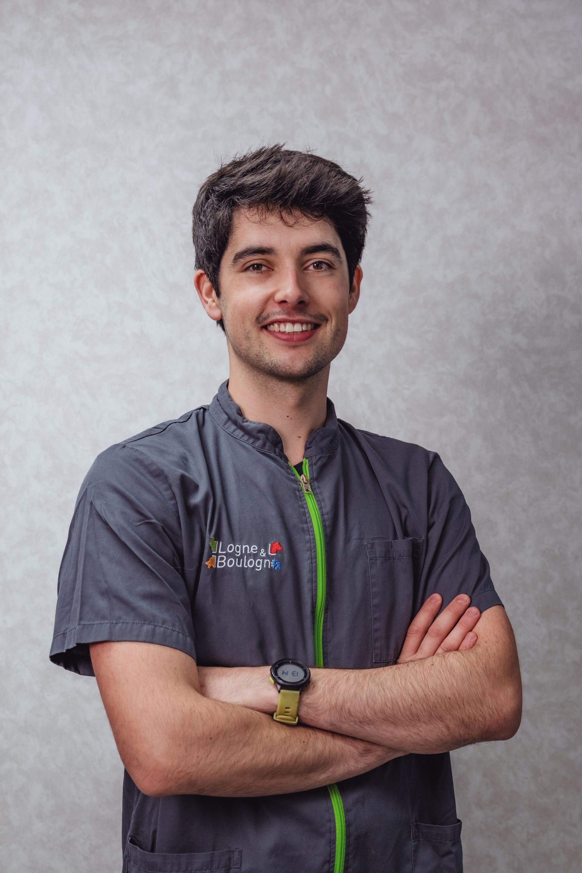 Un homme en blouse grise sourit, les bras croisés, devant un fond gris.