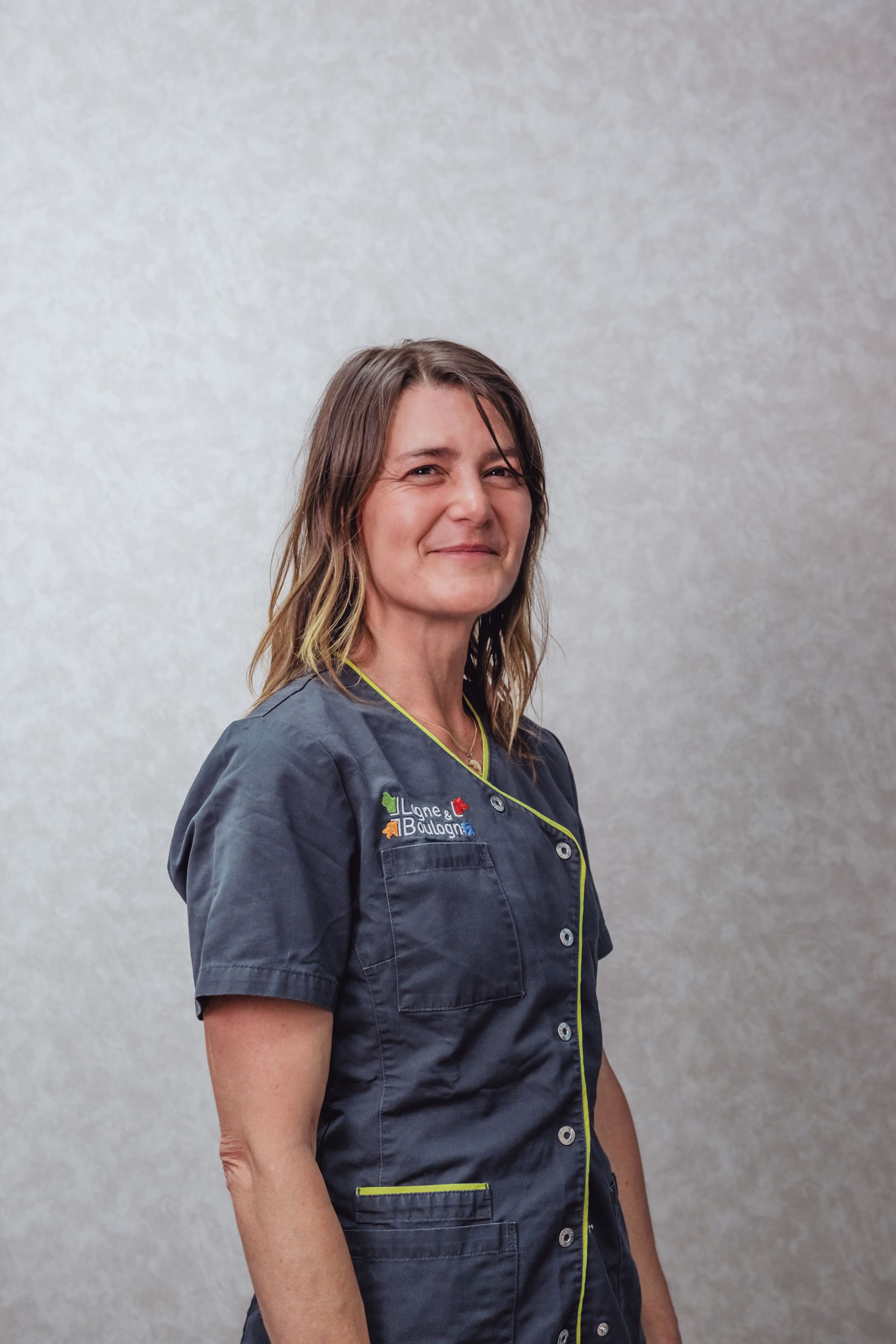 Une femme en blouse bleue sourit devant un fond neutre.