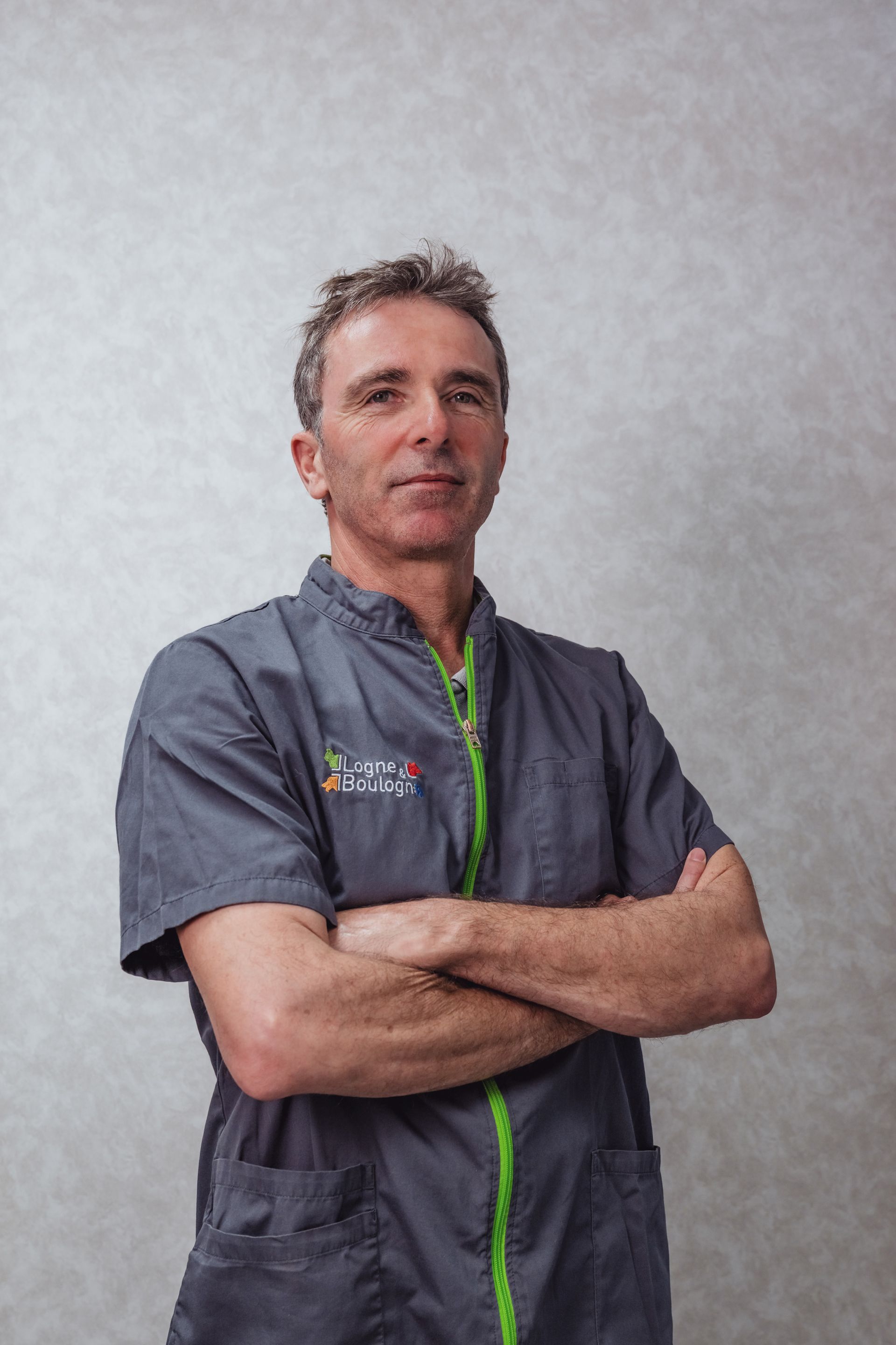Un homme en blouse médicale grise, les bras croisés, fermeture éclair et logo verts, se tient devant un mur gris clair.