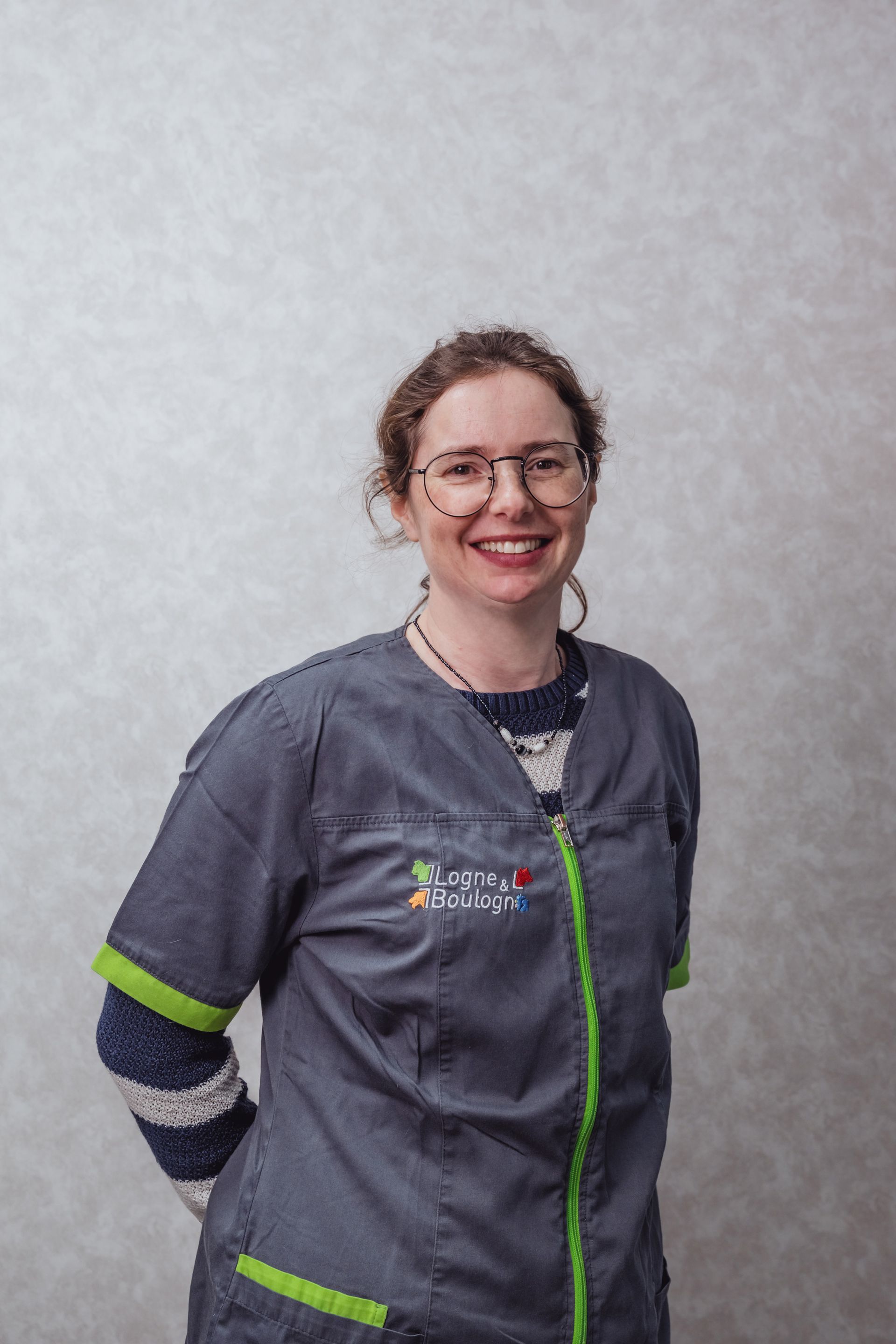 Femme portant des lunettes et une blouse médicale, souriante, debout devant un mur gris texturé.